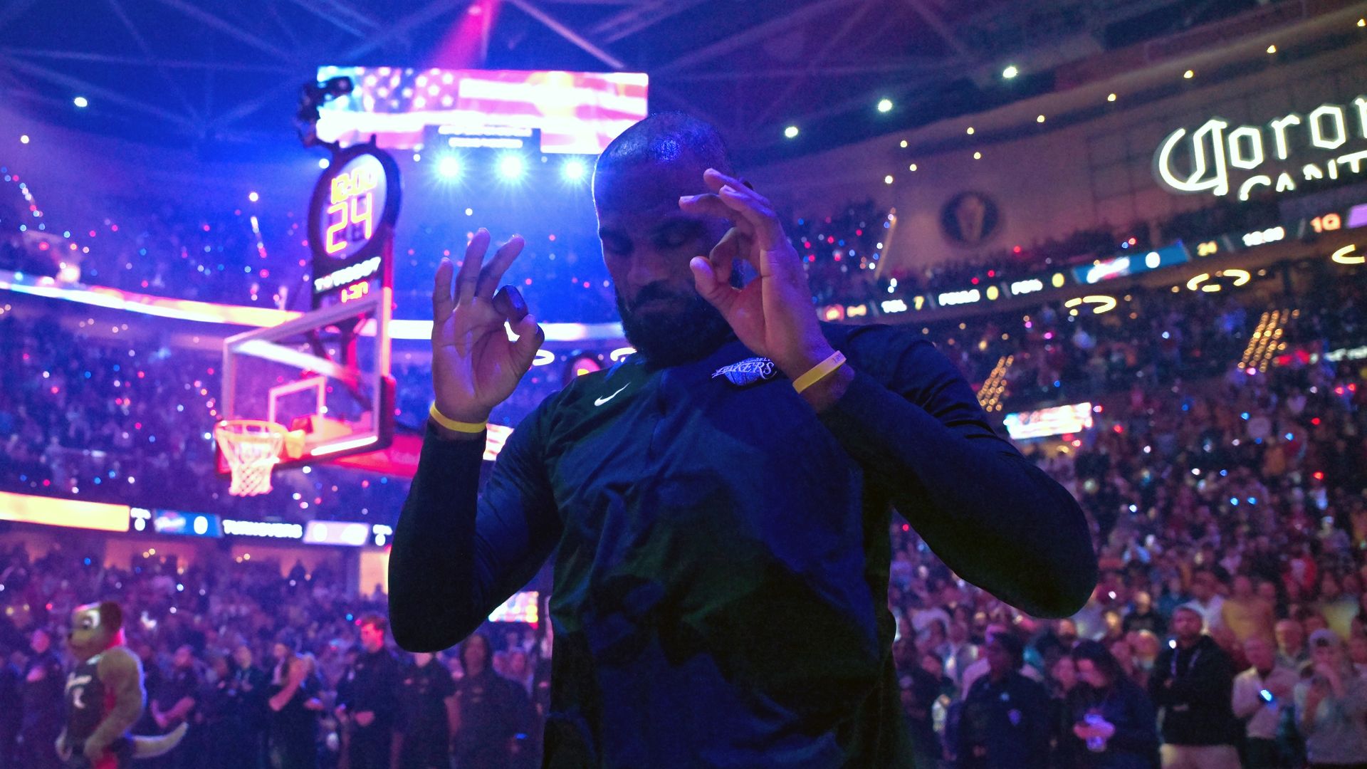 Inside a basketball arena lit in purple and pink, a player in dark blue practice gear faces the camera, making an "OK" gesture with both hands. Teammates warm up, fans watch, scoreboard above.