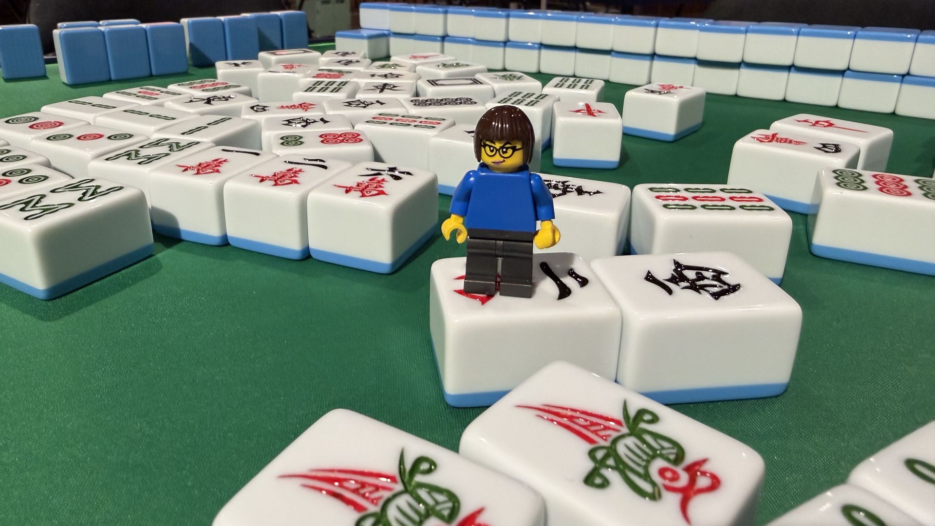A LEGO minifigure of Ina Fried in a blue shirt stands on a green Mahjong table, among white tiles with red and black symbols; blue tile racks and a blurred room with people in the background.