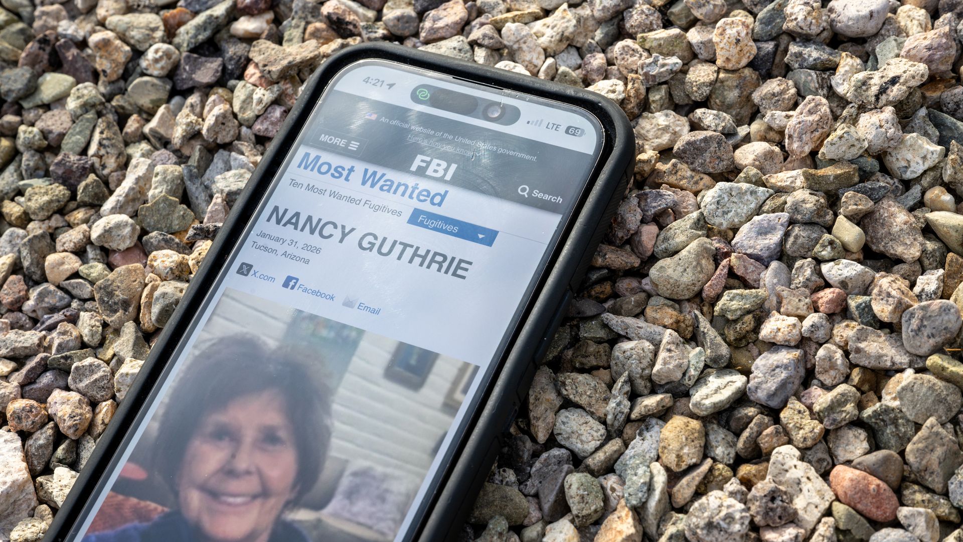Phone screen showing Nancy Guthrie’s profile on the FBI site with blurred Tucson street background.