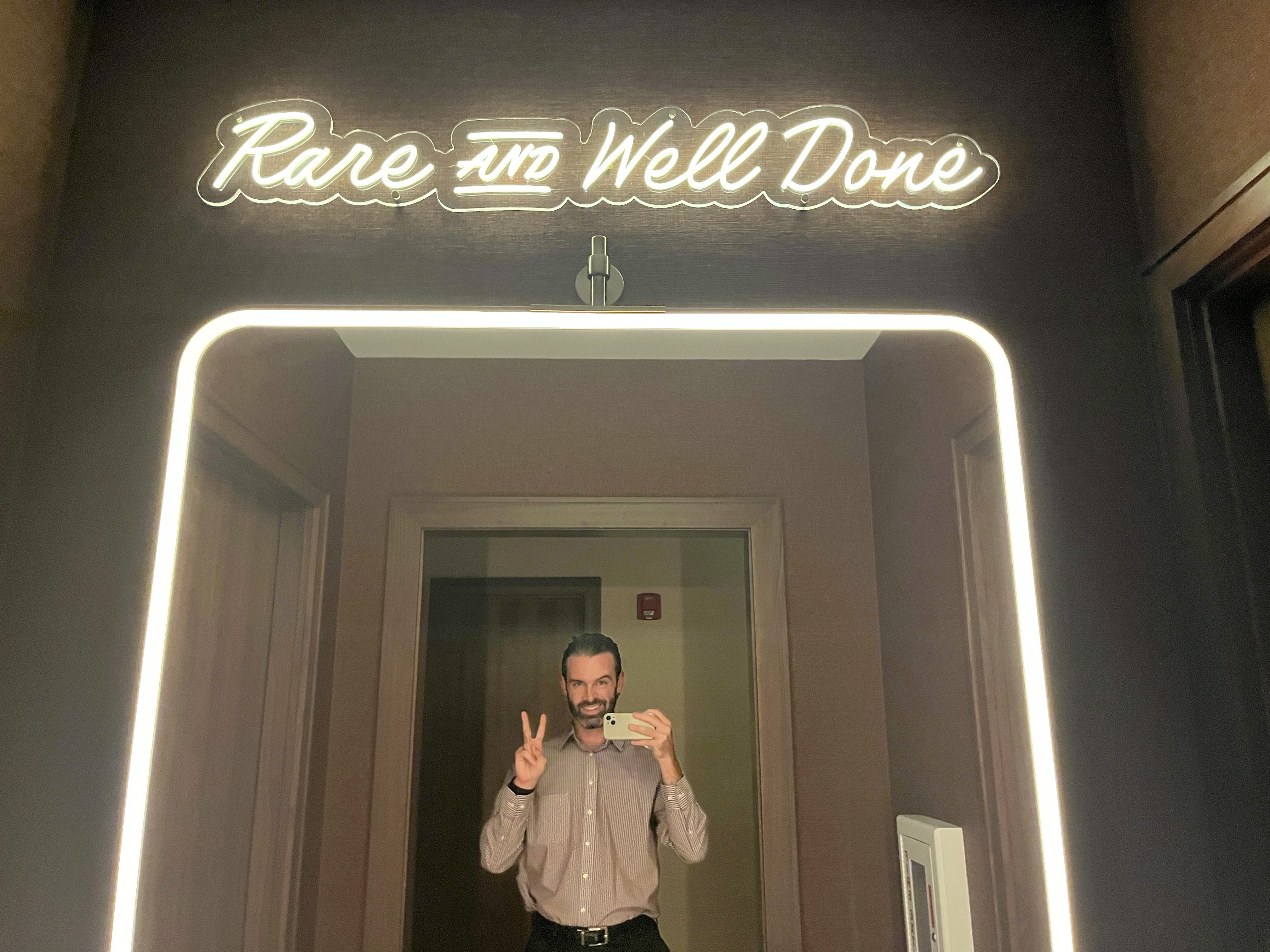 Man with dark hair and beard in a light pink shirt taking a mirror selfie and making a peace sign under a neon sign that reads "Rare and Well Done" in a room with brown walls.