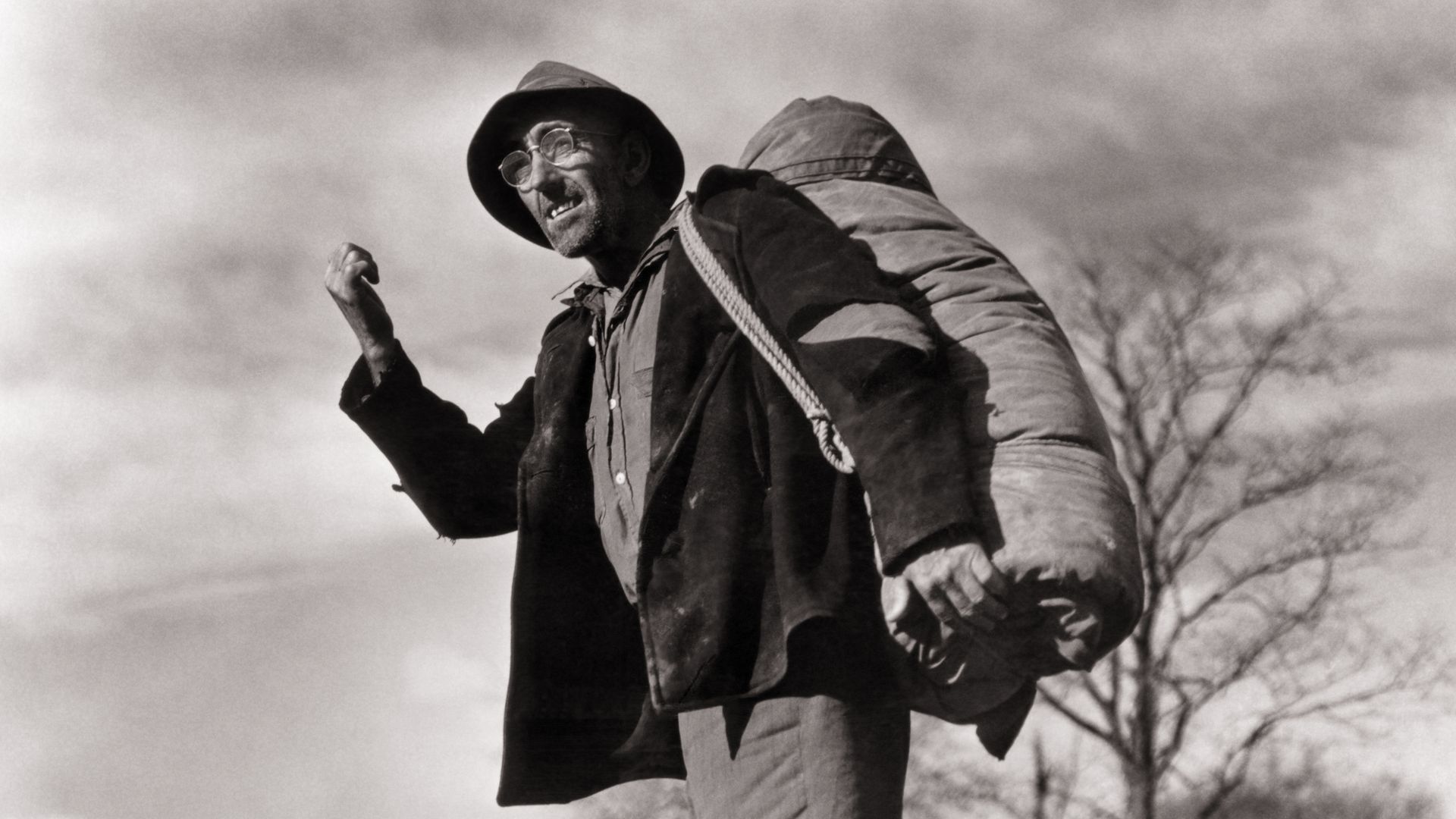 Black-and-white photo of a man standing by a rural road, wearing a hat and glasses, with a large sack over his shoulder, raising one arm as if hitchhiking.