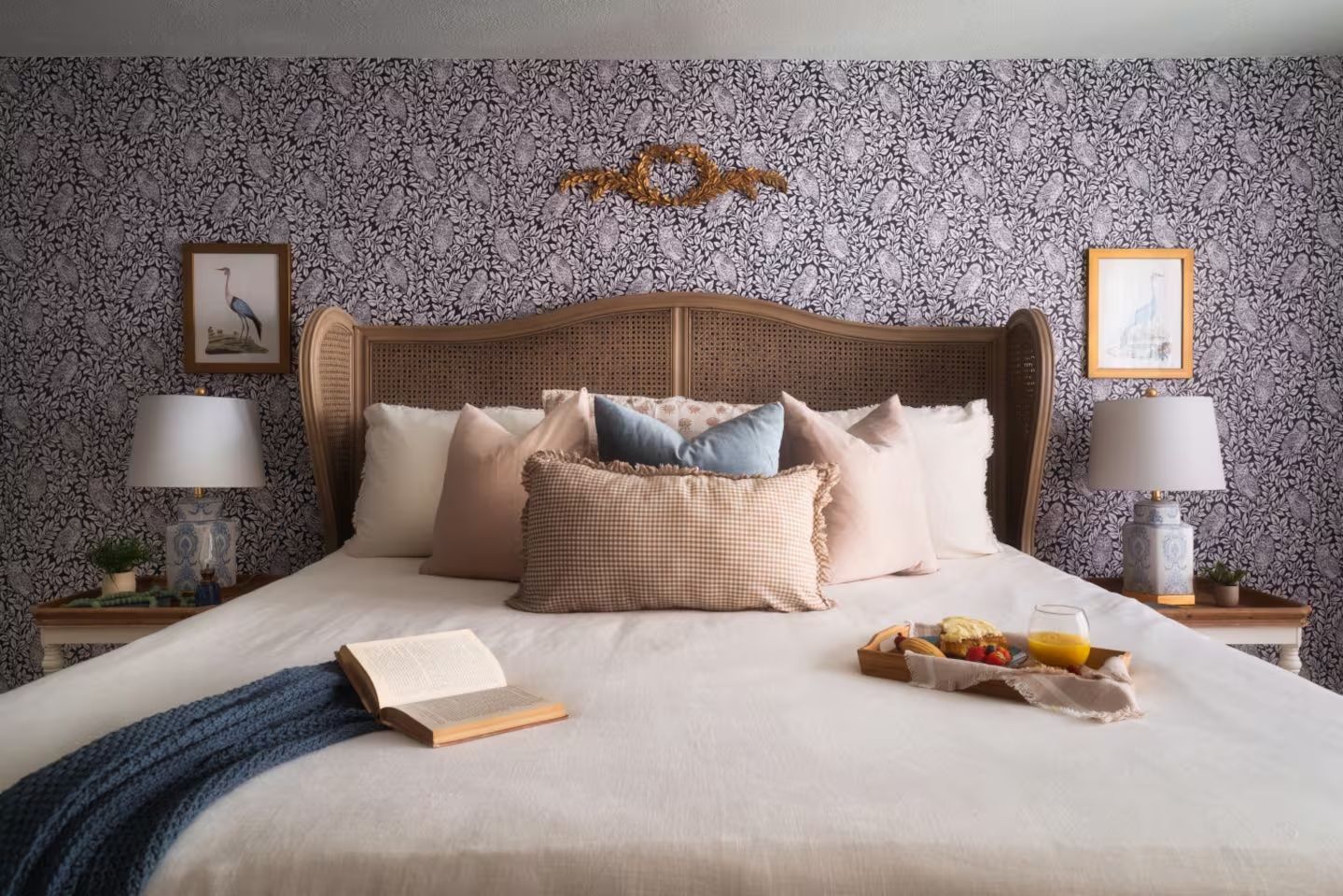 Cozy bedroom with patterned dark wallpaper, wooden headboard, multiple pillows in beige, blue, and pink, white bedding, open book, blue knitted throw, and breakfast tray with orange juice and pastry.