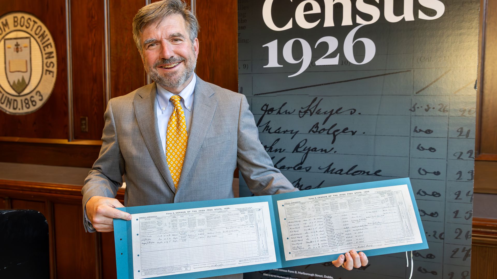 Boston College Provost David Quigley stands in front of a banner that says Census 1926 and holds up a folder containing copies of two census forms where each of his grandparents was recorded. 