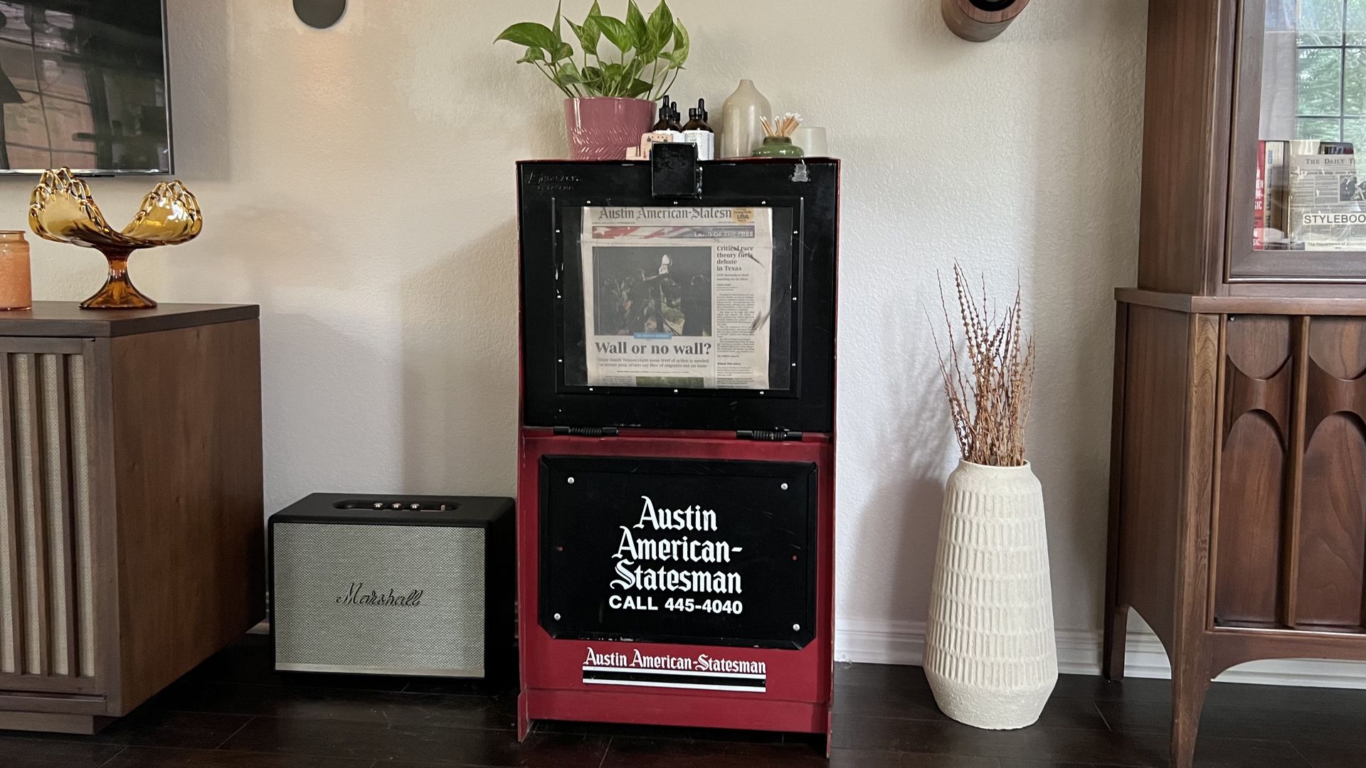Cozy living room with a red and black newspaper dispenser labeled "Austin American-Statesman" on the floor, topped by a green plant in a pink pot; amber bowl, lamp, guitar decor, and a white vase.