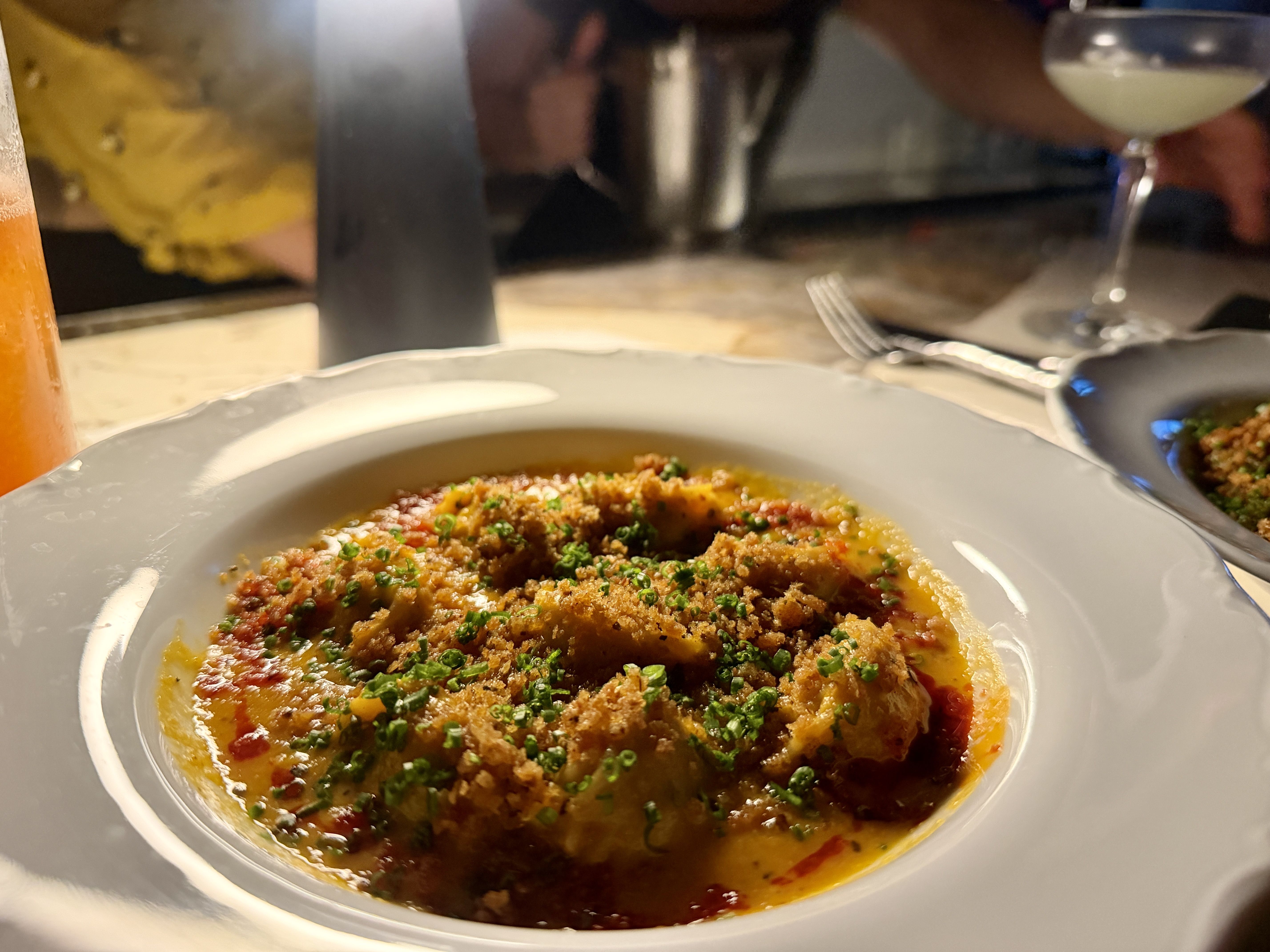 Close-up of a white bowl of yellow-orange soup topped with breadcrumbs and chopped chives. A fork lies nearby; blurred glassware and diners appear in the background.