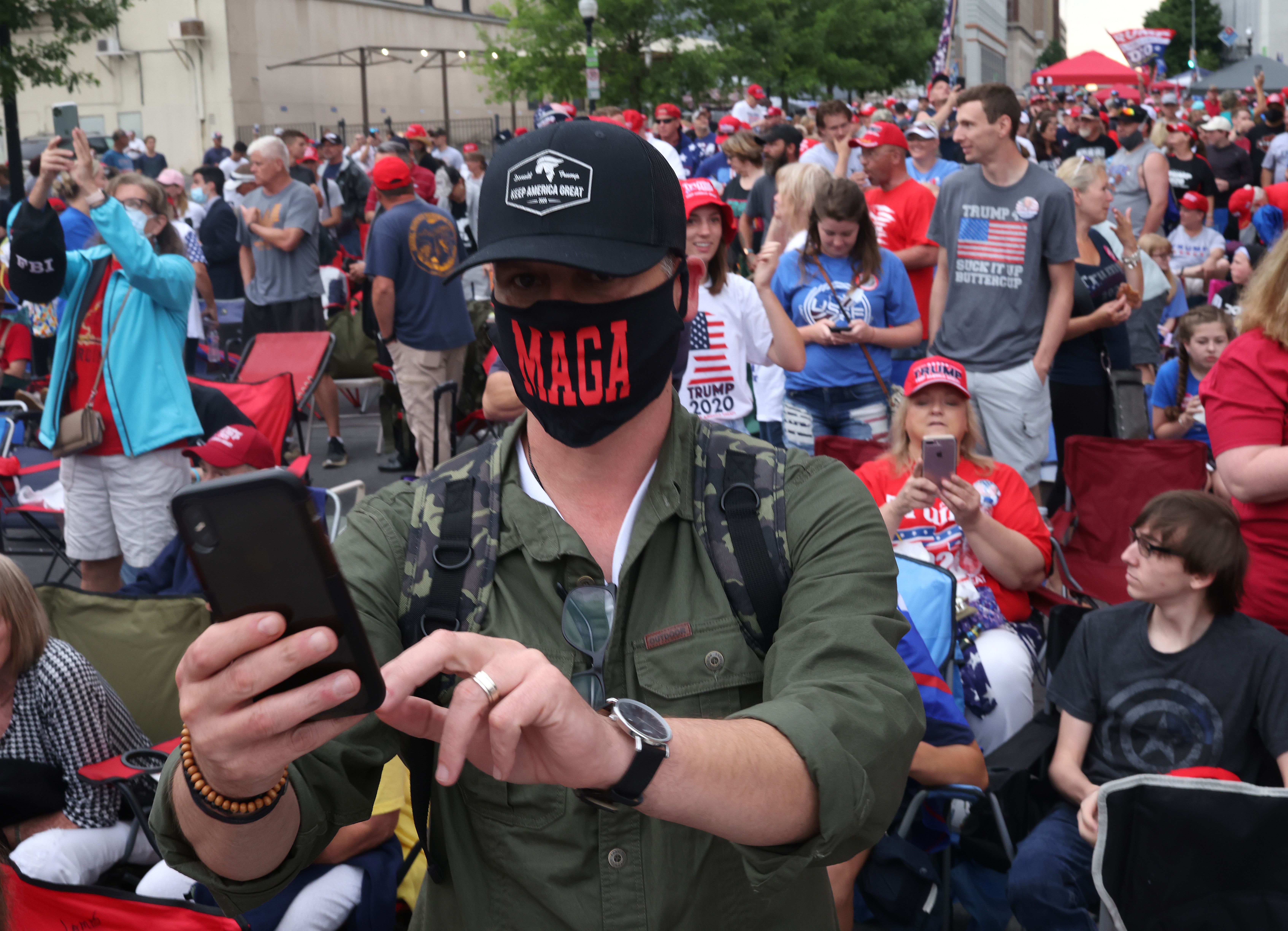 A man wearing a MAGA face masks holds a phone in front of a crowd of people