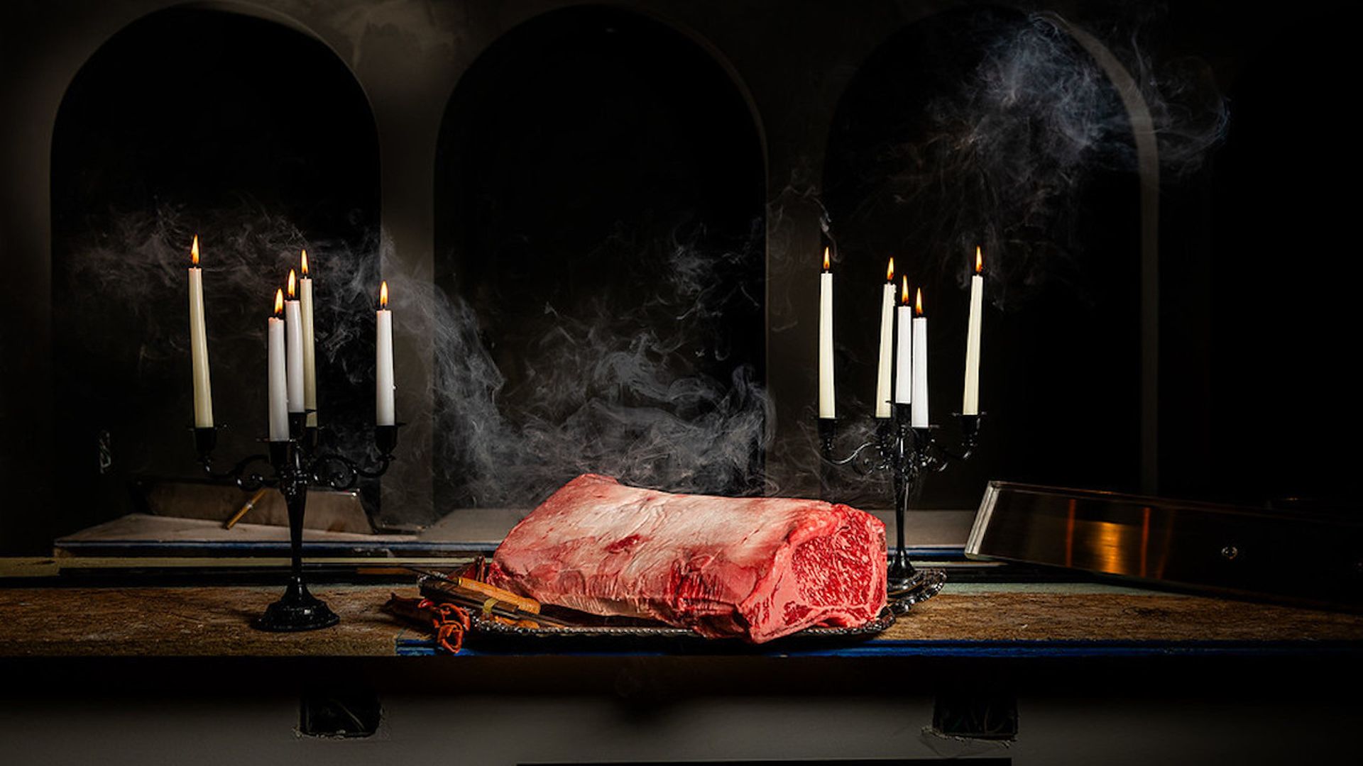 Dimly lit scene with a large raw beef roast on a platter, flanked by two black candleholders with lit candles; wisps of smoke rise around the meat on a dark table.
