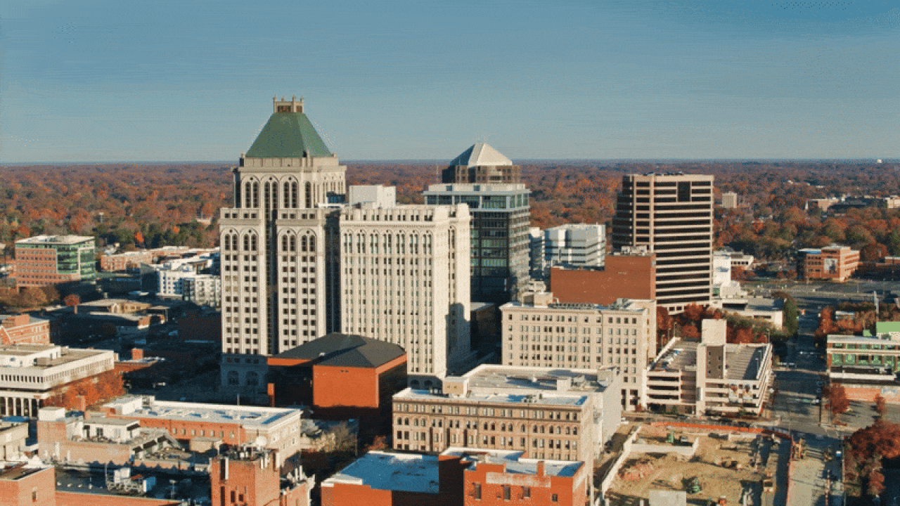 Aerial view of downtown Greensboro, North Carolina, showing a cluster of mid-rise and high-rise buildings surrounded by trees with autumn foliage under a clear blue sky.