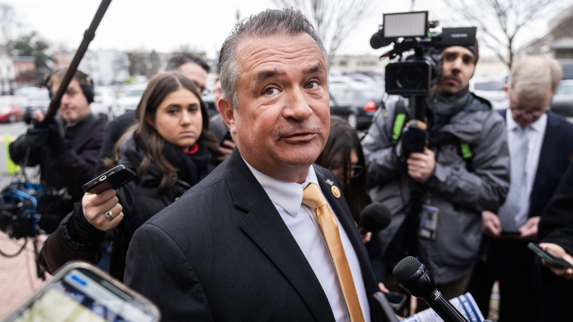 Rep. Don Bacon, wearing a dark gray suit, light blue shirt and gold tie, standing outside surrounded by press.