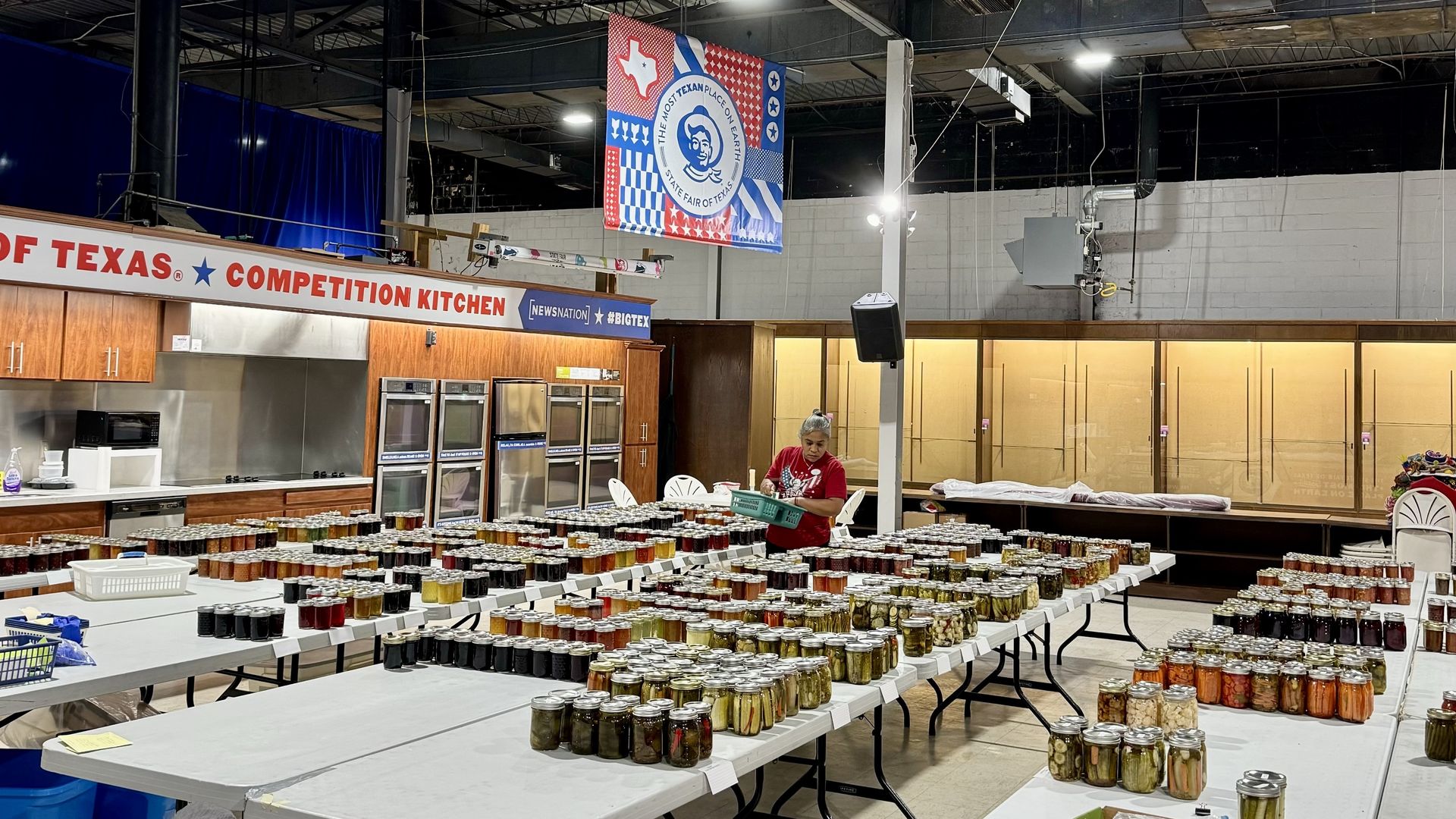 Indoor competition kitchen with tables covered in hundreds of glass jars of pickled vegetables and jams, a person arranging jars, and a colorful Texas State Fair banner overhead.