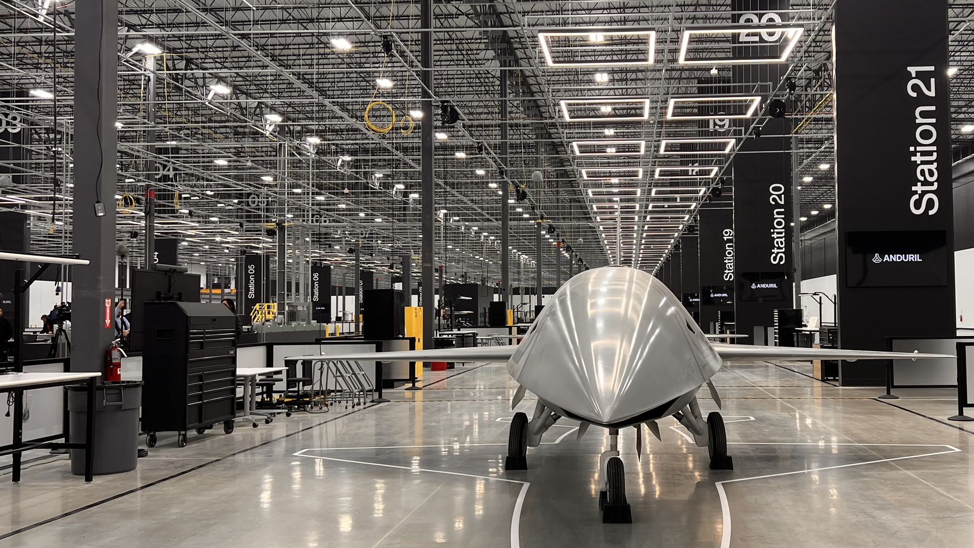 Inside a large aircraft hangar, a sleek silver triangular aircraft on wheels sits center. Bright overhead lights, station signs (Station 20/21), and a man in a jacket taking photos nearby.