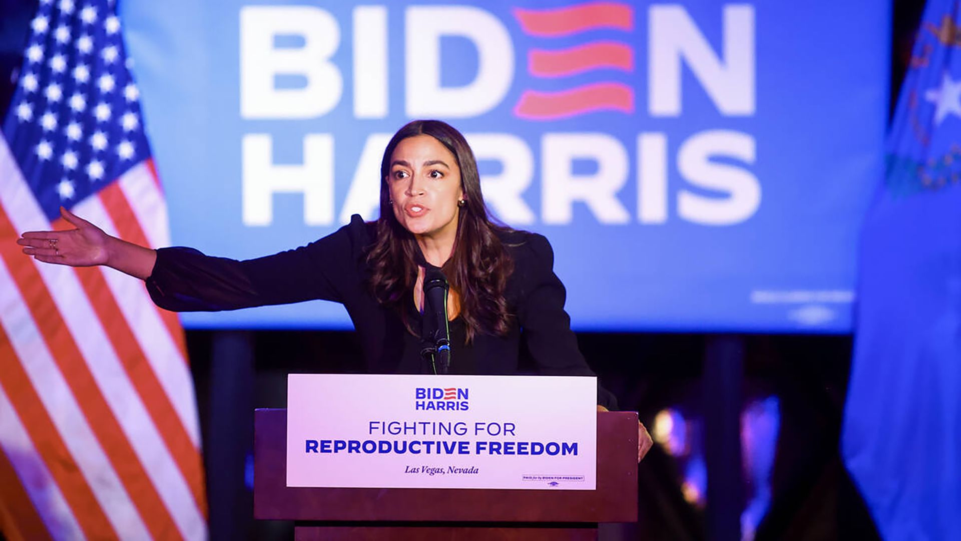 Rep. Alexandria Ocasio-Cortez speaking at a podium in front of a blue Biden-Harris sign.