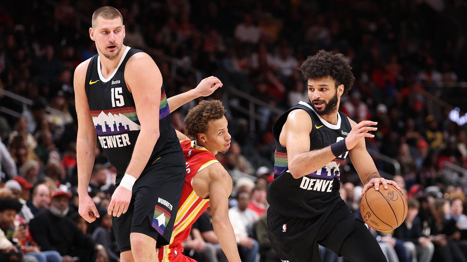 Denver Nuggets players in black rainbow-themed jerseys playing basketball against a team in red and yellow uniforms, with one player dribbling the ball on the court during a game.