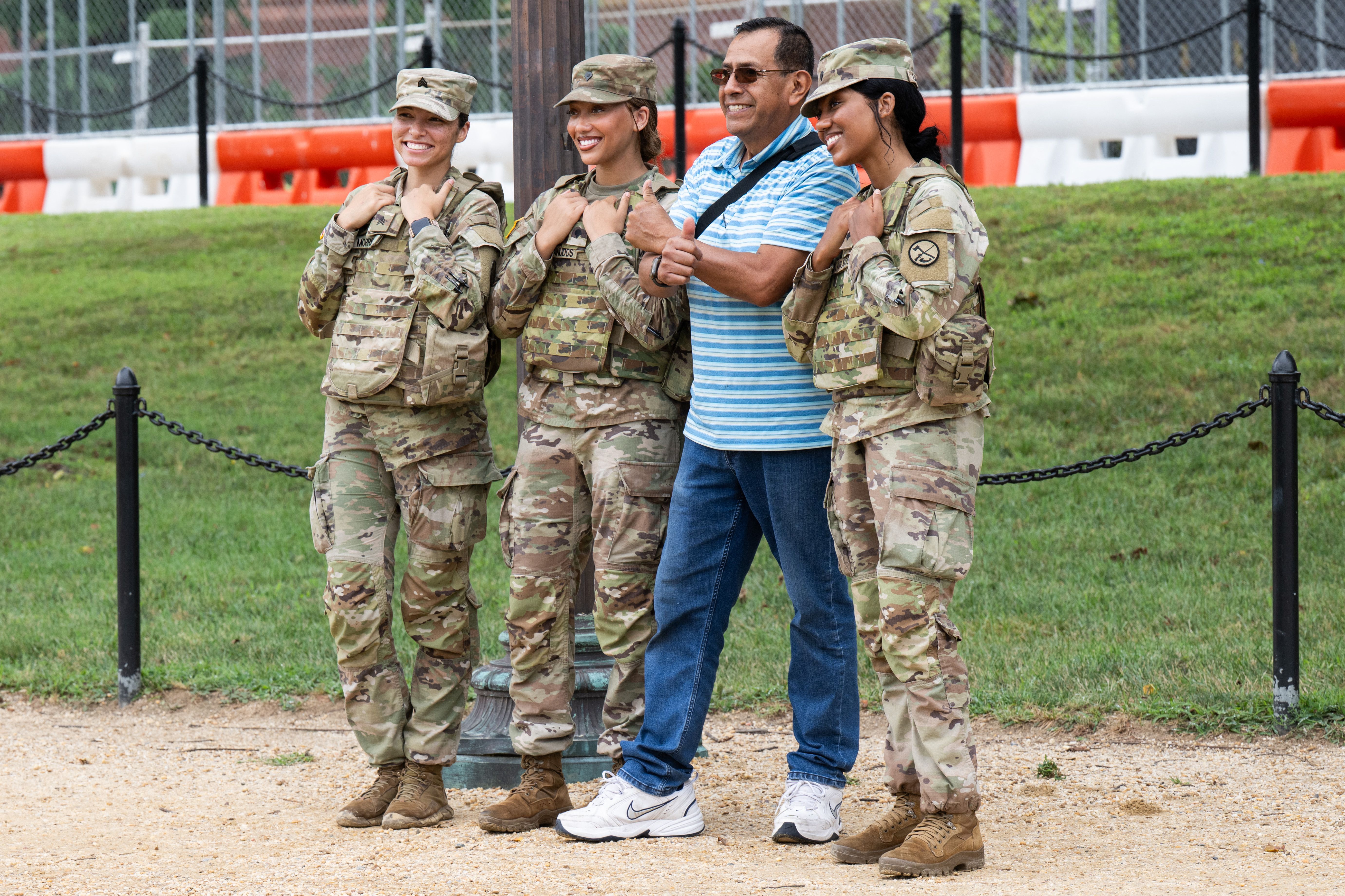 Three women in camouflage military uniforms and a man in a blue striped shirt and jeans stand smiling outdoors on gravel with grass and orange-white barriers behind.