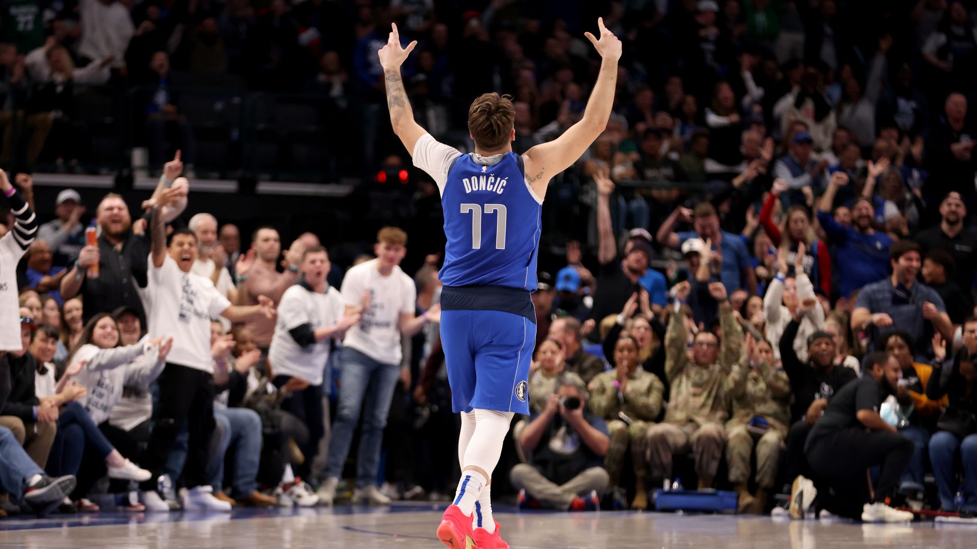 A basketball player in a blue uniform faces the crowd in an arena with his arms up