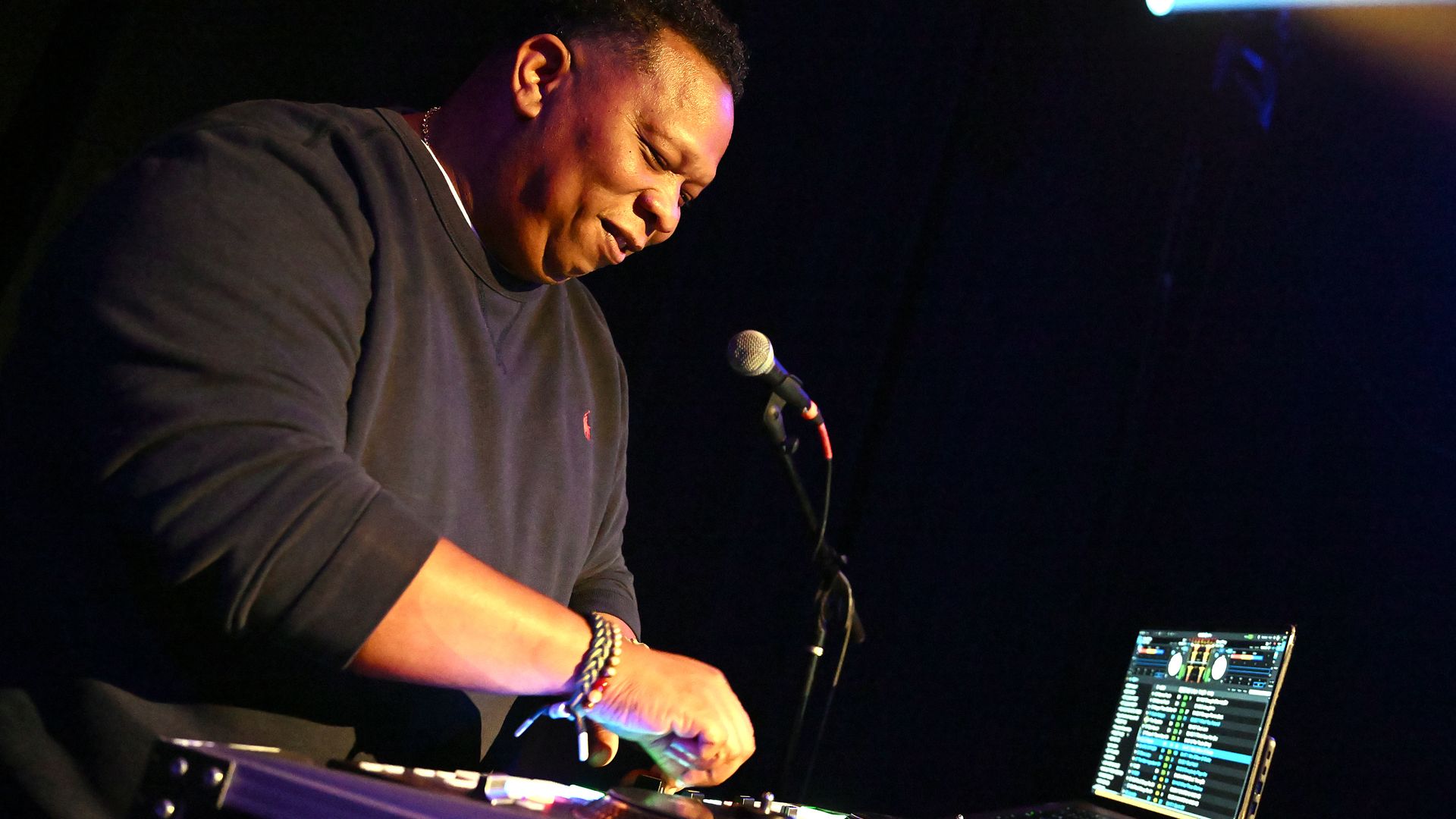 Mannie Fresh smiles as he leans over a mixing board and a microphone with a laptop open in the background.