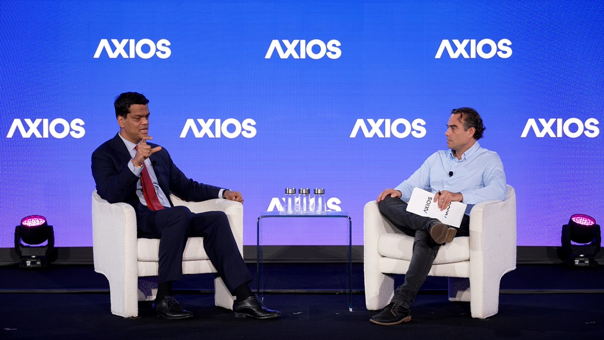Two men seated in white chairs on a stage with multiple "AXIOS" logos on a blue background. The man on left wears a dark suit, red tie; the man on right wears light blue shirt, holds "AXIOS" paper.