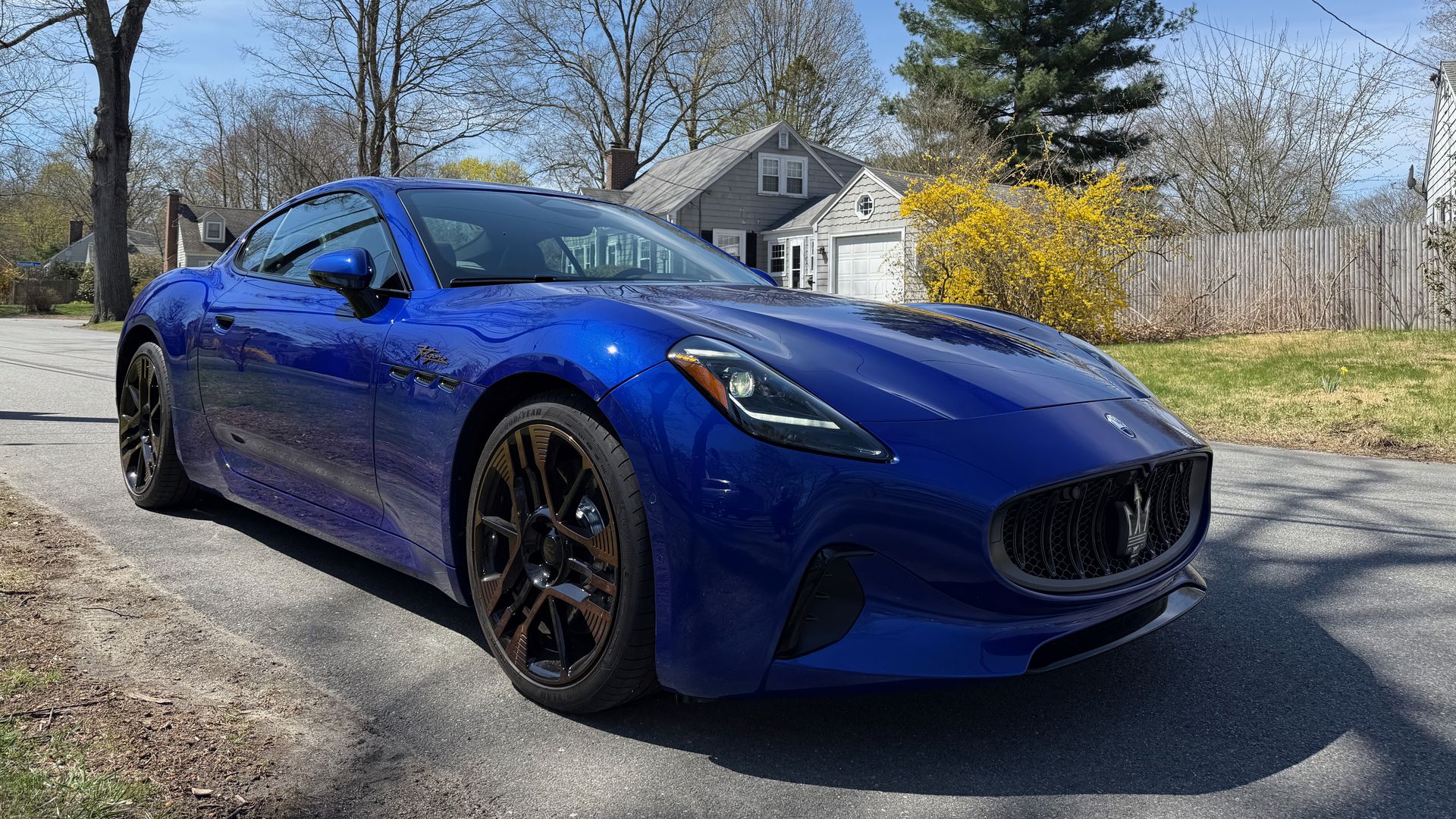 A bright blue Maserati Folgore Gran Turismo with dark bronze wheels parked on a suburban street. In the background, leafless trees, a yellow-flowering bush, a gray house, and a wooden fence under a clear blue sky.