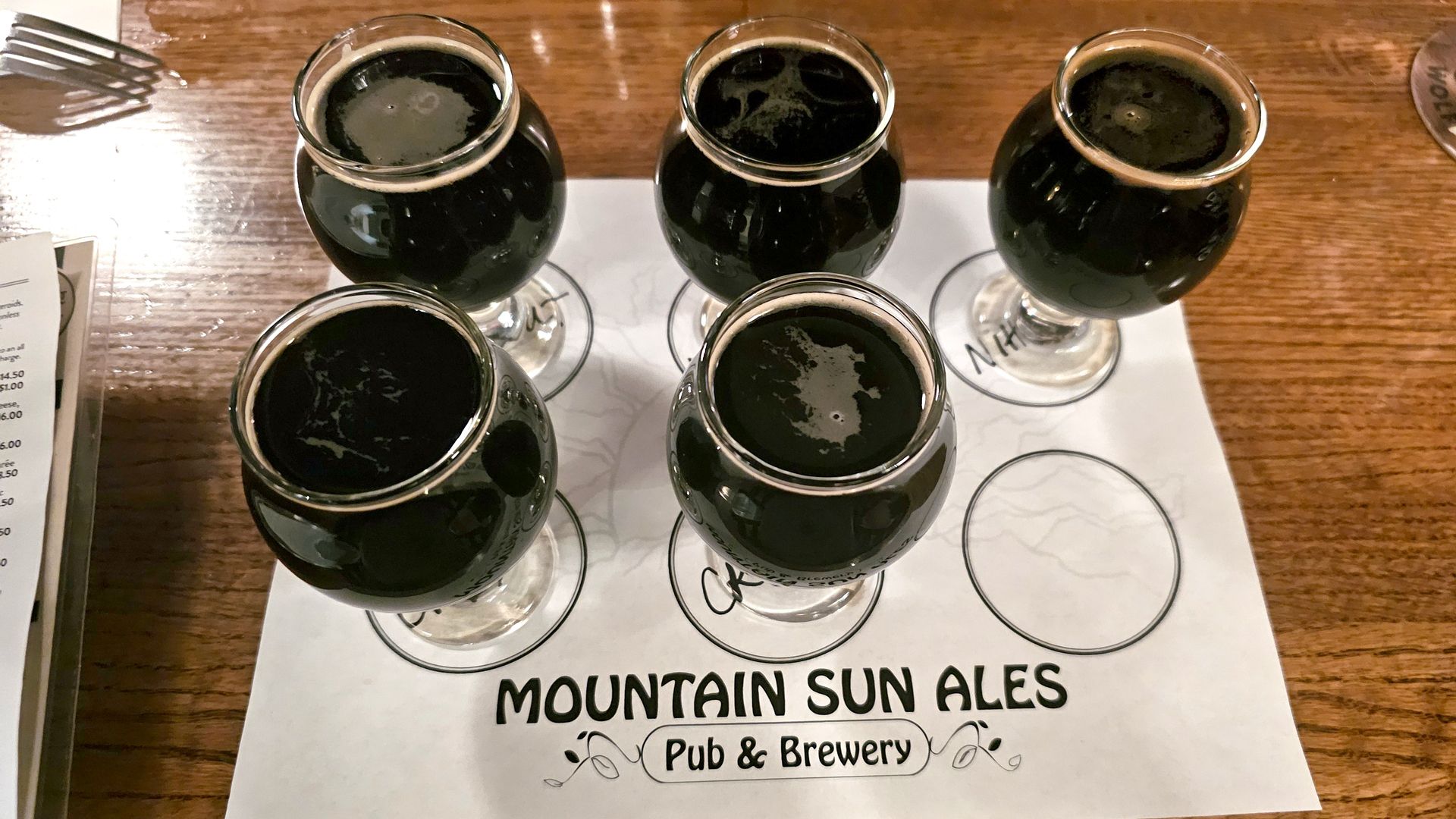Five small glasses of dark beer arranged on a Mountain Sun Ales Pub & Brewery tasting mat on a wooden table.