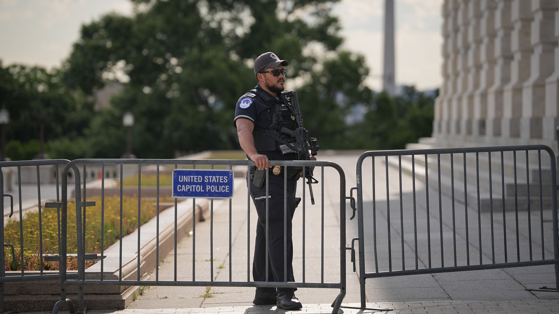 A Capitol Police officer wearing a black uniform, hat and sunglasses stands behind bike rack fencing in front of a marble building and the Washington monument.