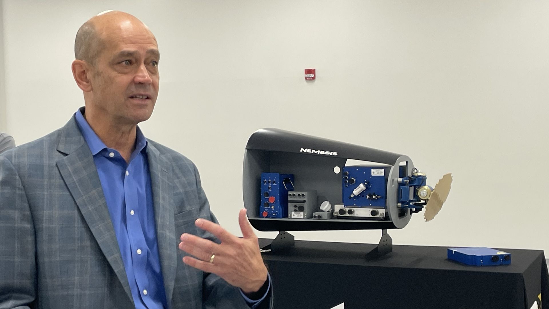 A bald man in a blue shirt and blue plaid blazer speaks, gesturing with his hands. In the background, another man in a gray polo watches. A blue mechanical device sits on a table beneath a large banner.