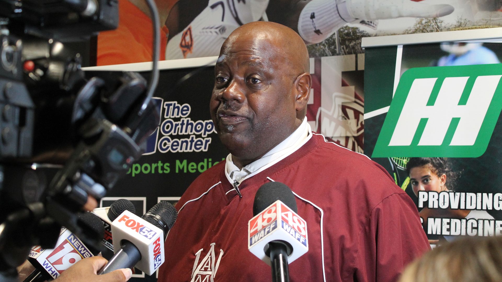 Man in maroon Alabama A&M Bulldogs sweatshirt being interviewed by multiple news reporters with microphones from Fox54, WAFF48, and News19, in front of sports medicine posters.
