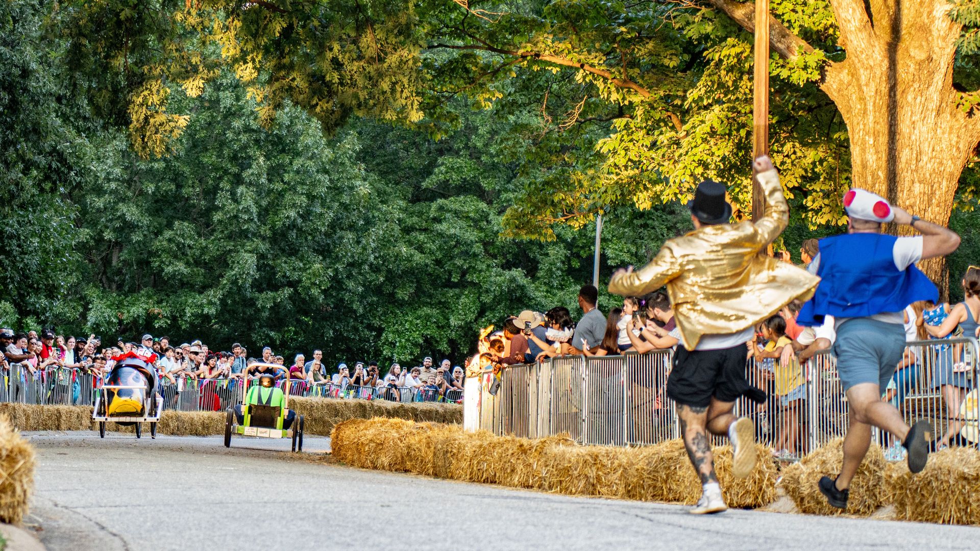 Crowded street event with two small race carts and cheering spectators behind barriers; gold-clad performers sprint beside hay bales as green trees tower overhead.