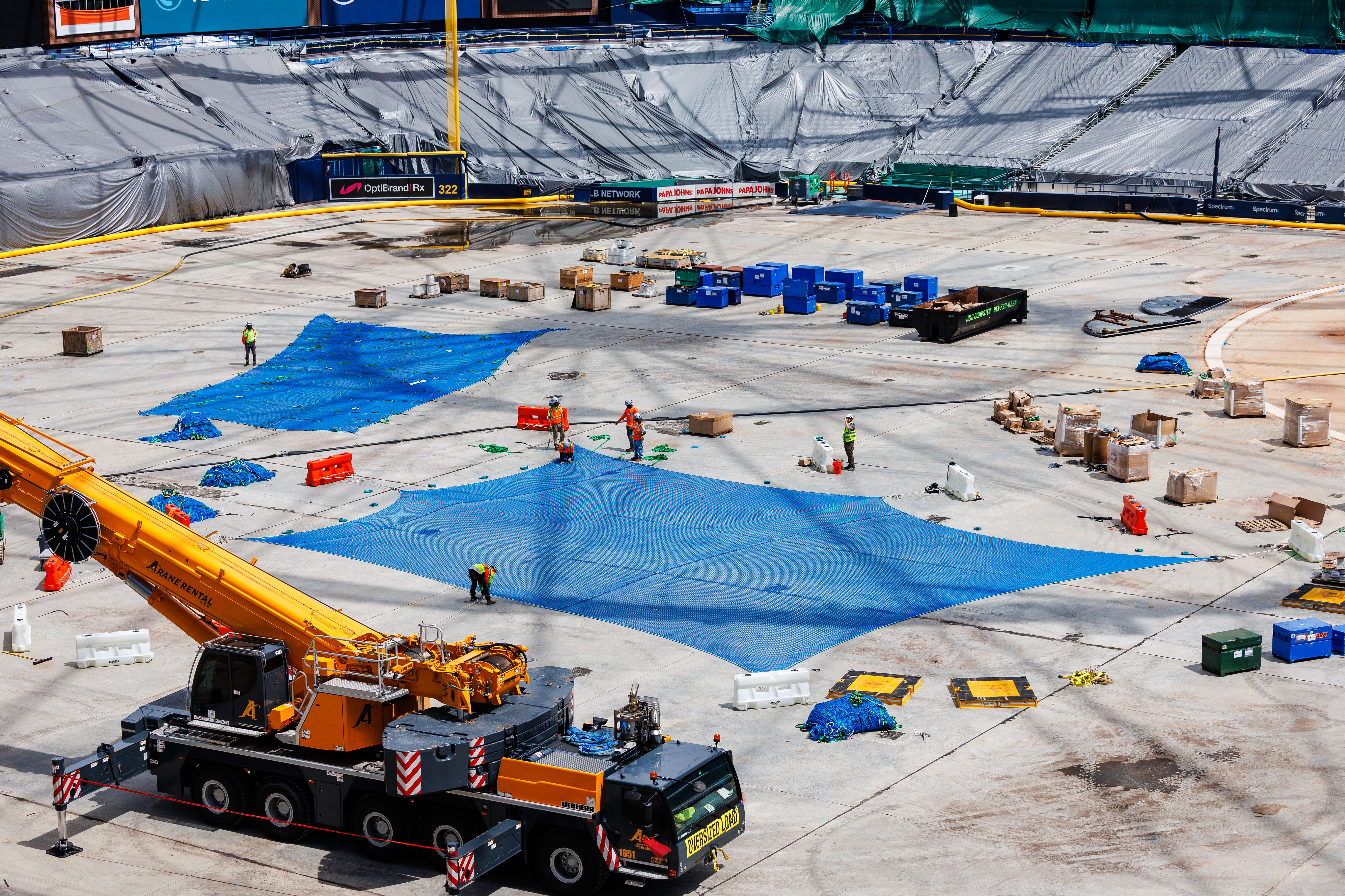 Construction site with workers in orange vests, blue nets spread on the ground, a yellow crane truck and scattered crates and equipment on concrete surface.