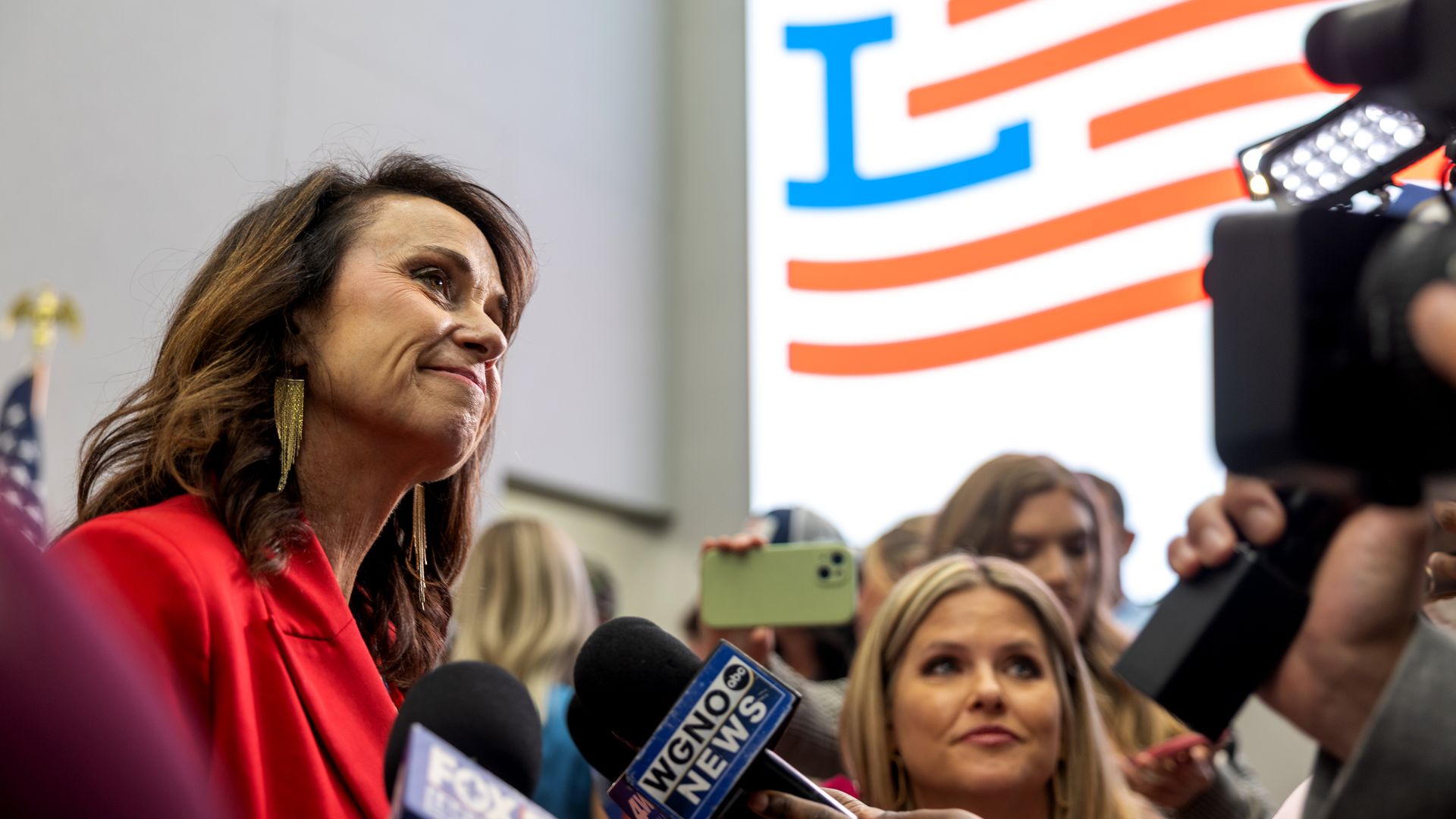 Image shows Liz Murrill, in a red jacket, smiling at a TV camera with a microphone in front of her.