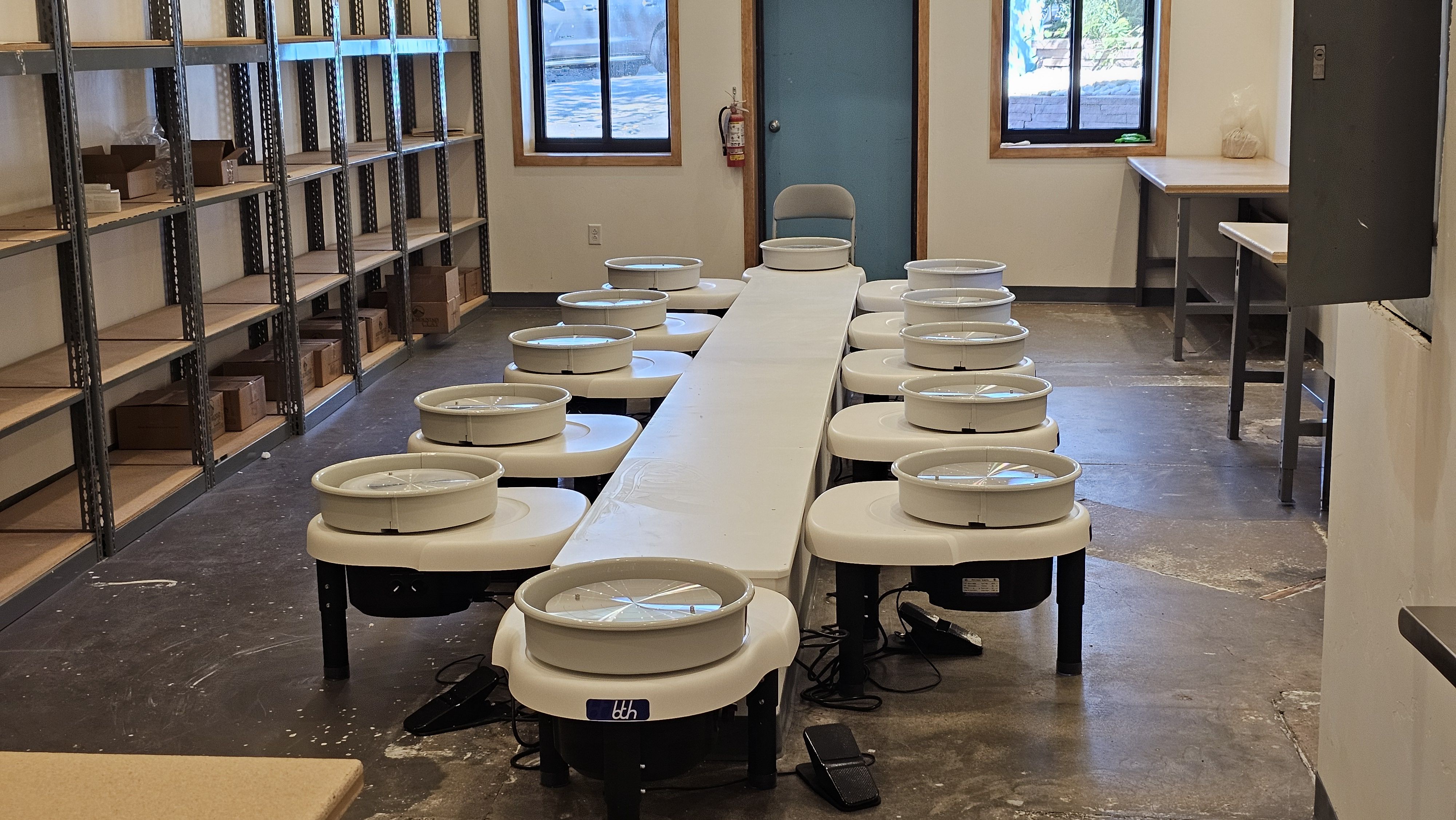 Industrial workshop with a long central table lined with white circular devices with lids, flanked by metal shelving filled with boxes; blue door, two windows, and a fire extinguisher on the wall.