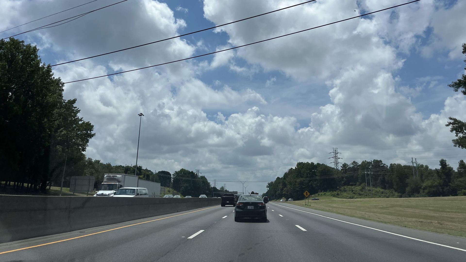 Roadway with several lanes of cars beneath a blue sky and fluffy clouds. Power lines cross the frame, with trees on either side and a concrete barrier left.