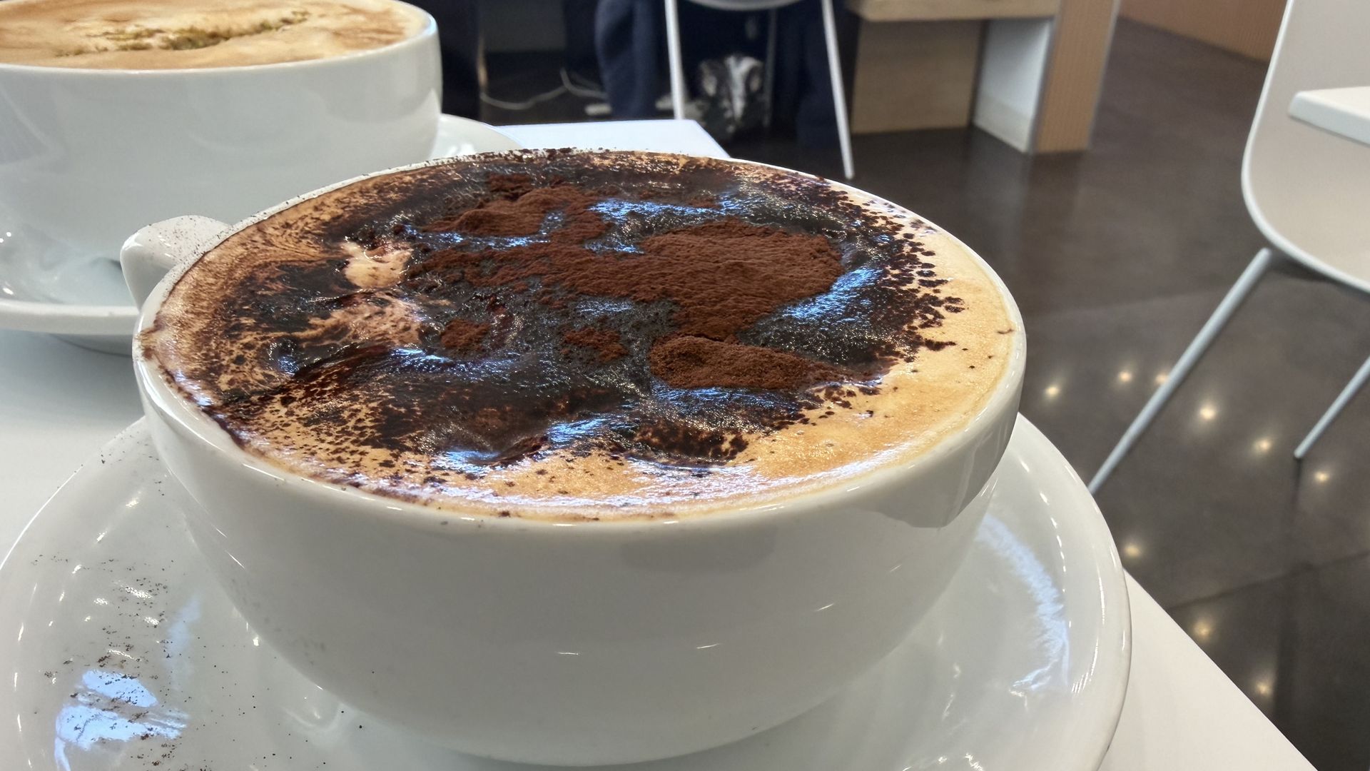Close-up of a white cup of cappuccino with frothy milk and cocoa powder on top, sitting on a white saucer at a cafe with modern chairs and shiny floor.