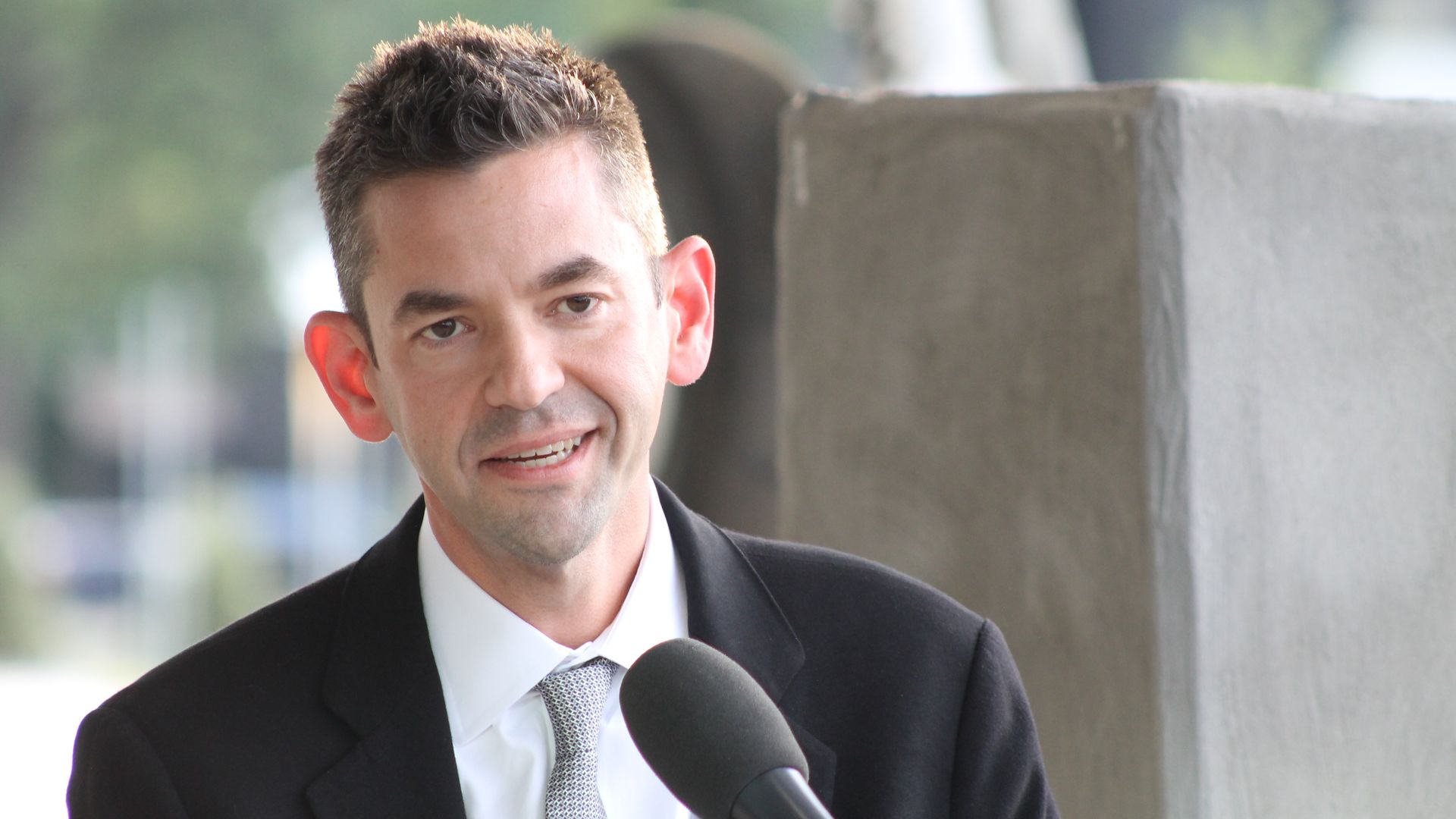 A man in a black suit and white shirt with a gray patterned tie speaks into a handheld microphone outdoors, with a blurred green background and concrete wall behind him.