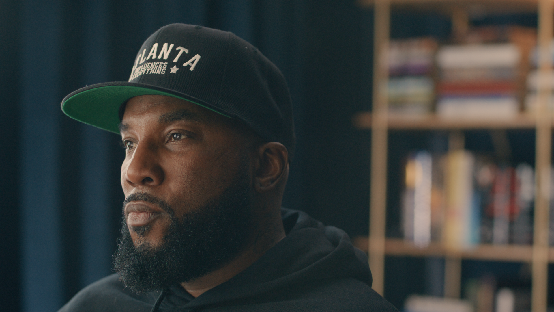 A photo showing a portrait of Jeezy wearing an Atlanta baseball hat and a hoodie sitting in front of a blurred out bookshelf. He’s looking serious and at something out of the frame. He has a beard. 