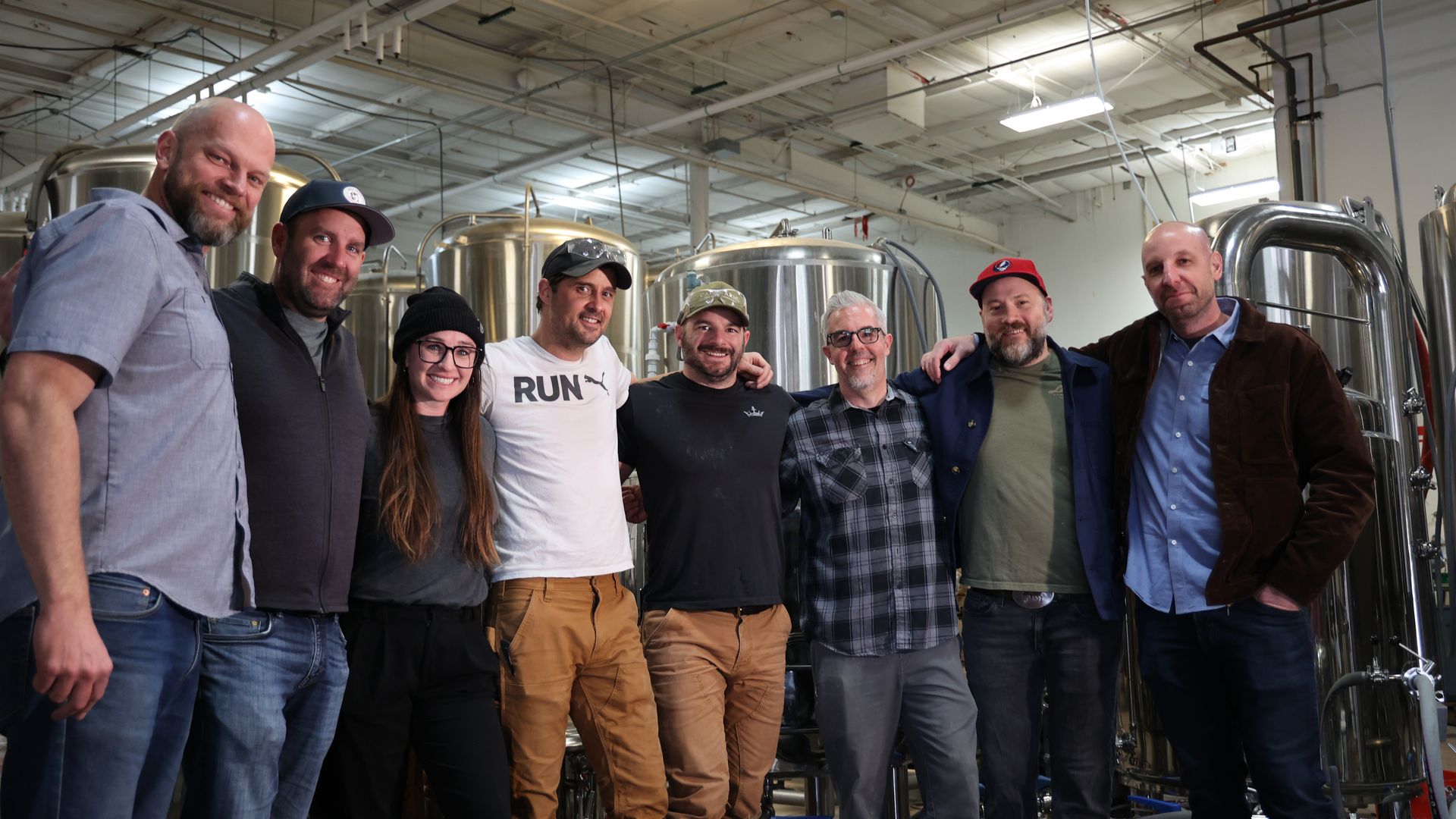 Group of nine adults smiling and posing in a brewery, standing in front of tall stainless steel fermentation tanks and exposed overhead pipes.