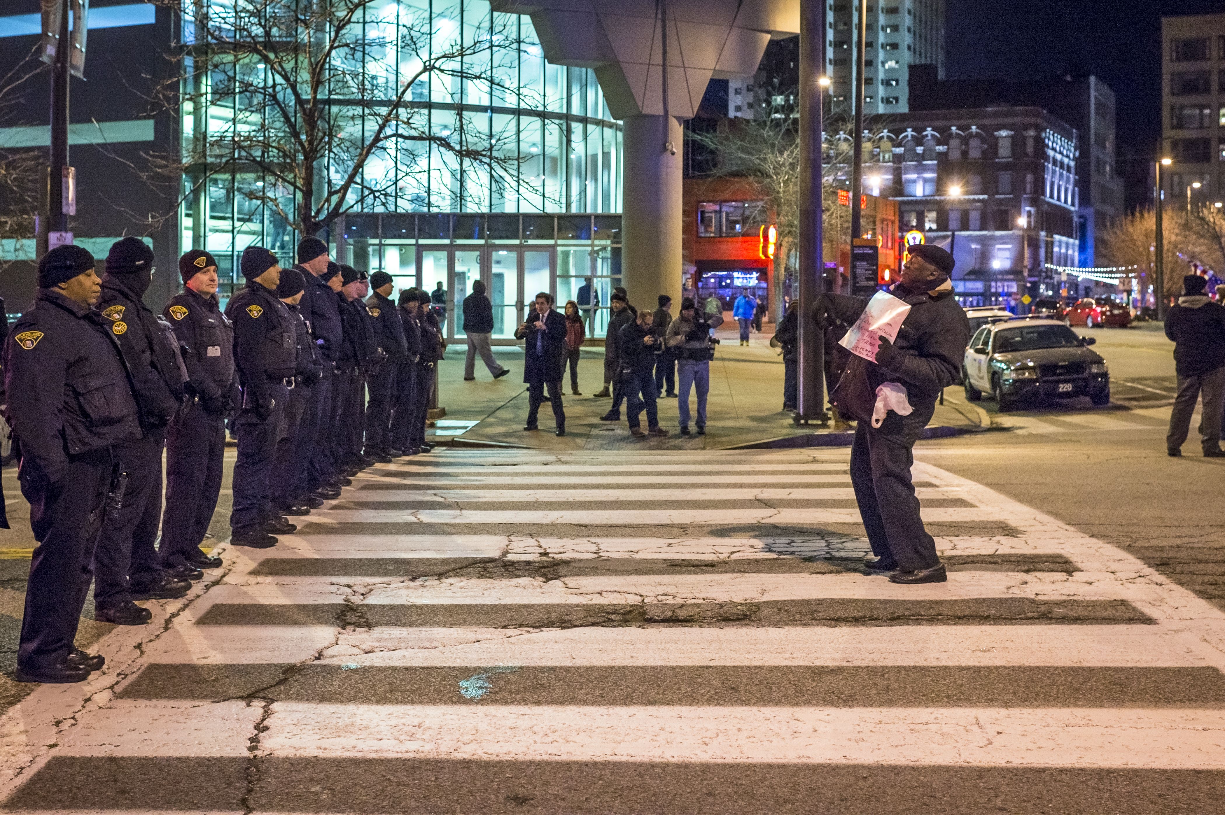 A defiant protester stands before a line of police officers at night at crosswalk