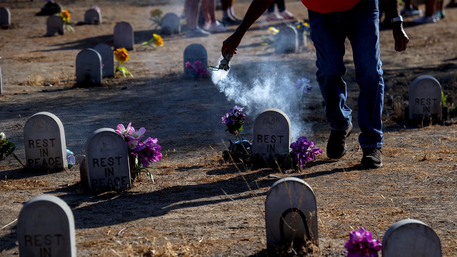 Photo of rows of graves sitting in brown dirt as a person encircles one with sage smoke