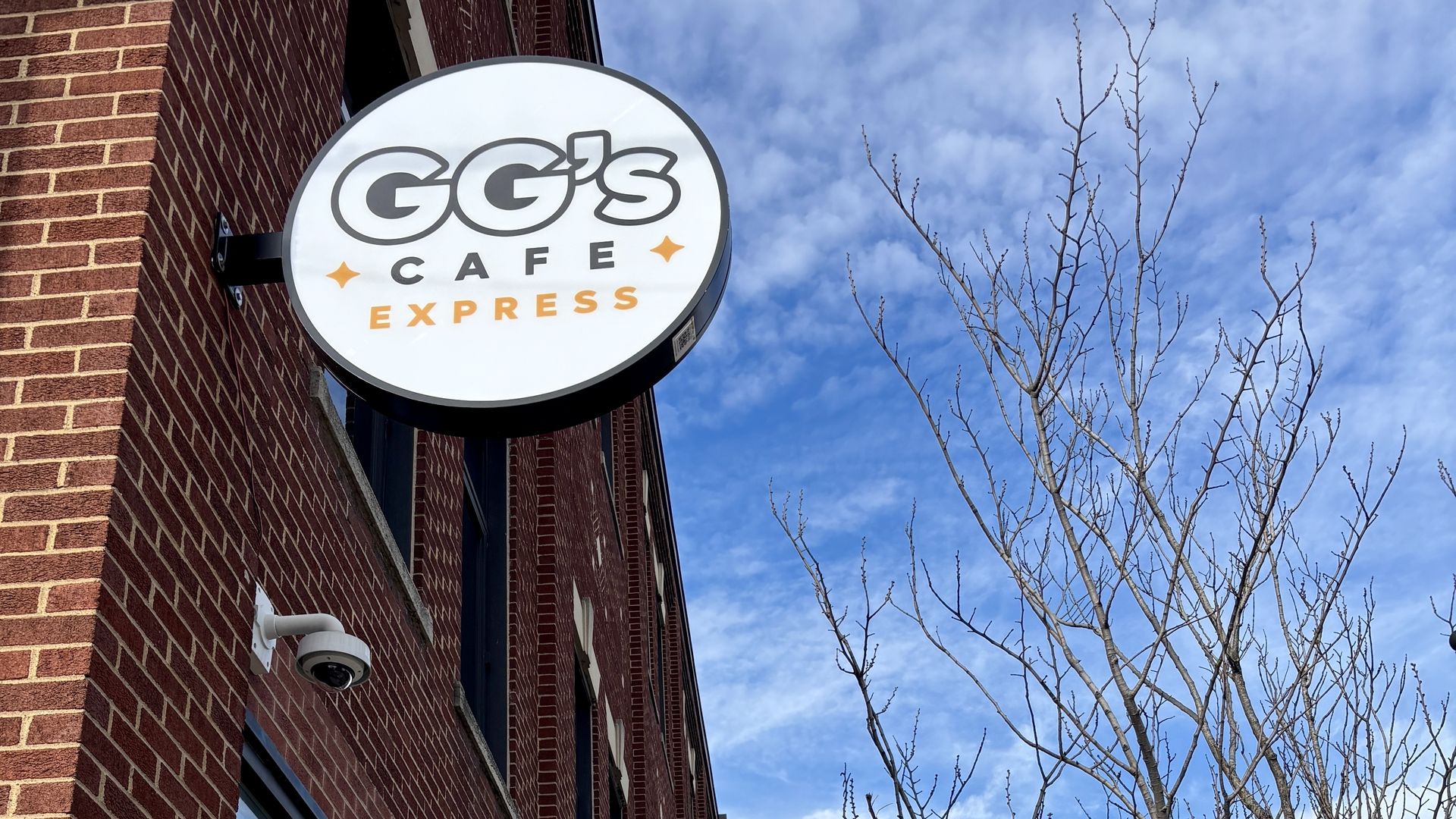 Round white sign on red brick building reading "GG's Cafe Express" in black and orange letters, with a blue sky and leafless tree branches in the background.