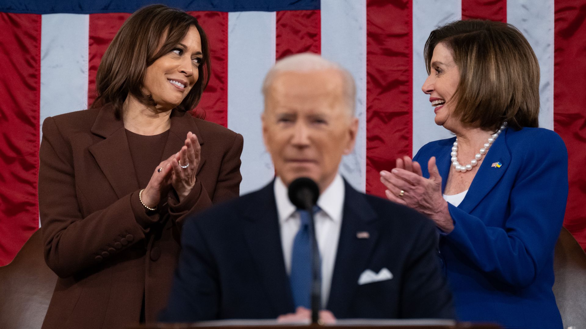 Kamala Harris wearing a brown suit and Nancy Pelosi wearing a blue suit, clapping in front of an American flag as Joe Biden speaks in the foreground.