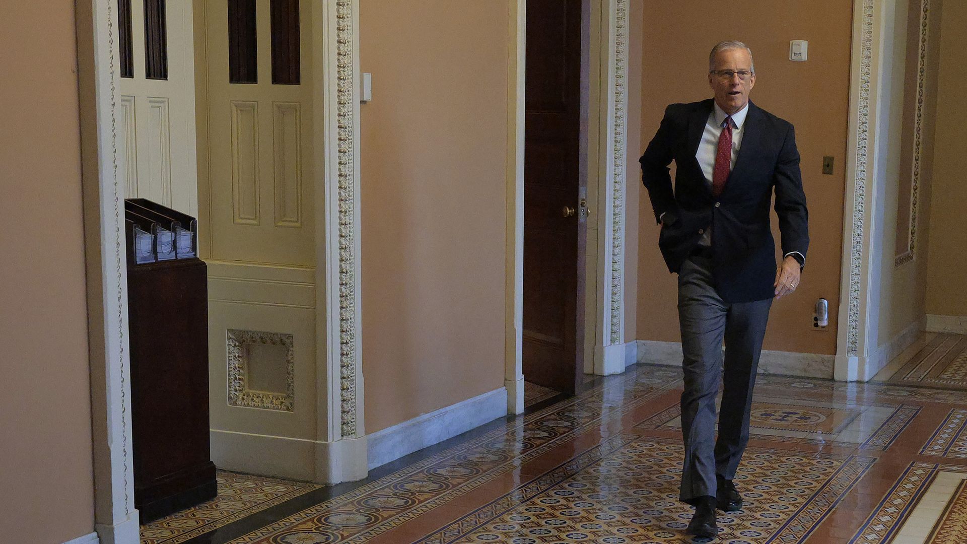 Man in dark suit, red tie, and glasses walking down ornate tiled hallway with peach-colored walls and white trim.
