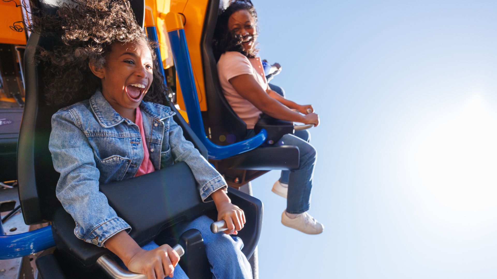 Two riders on a roller coaster: front girl with curly hair in a denim jacket screams, gripping a safety bar; behind her, a smiling girl in a pink shirt holds on as the bright blue sky shines.