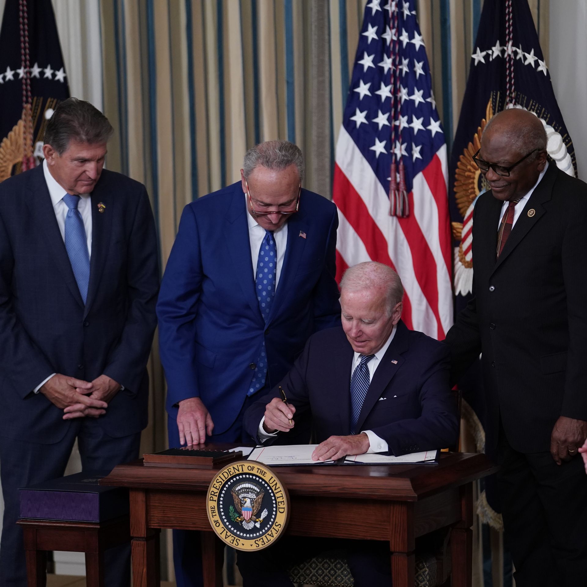 Photo of Joe Biden signing a bill on a desk with lawmakers crowded around and behind him