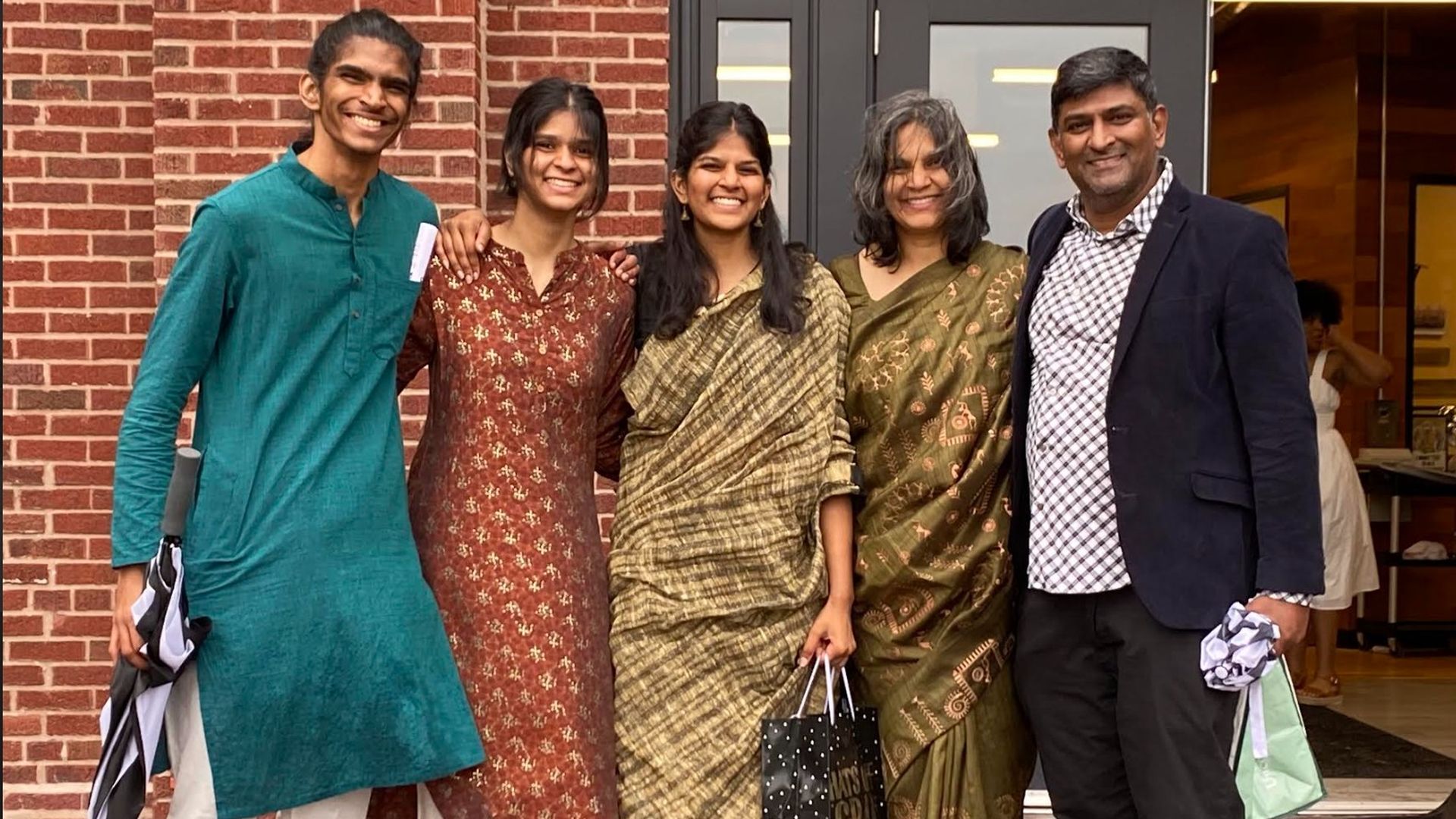 Five people stand outside a brick building, smiling. From left: a man in teal kurta, a woman in brown kurta, a woman in beige saree, a woman in olive saree, and a man in a blazer with a checkered shirt.