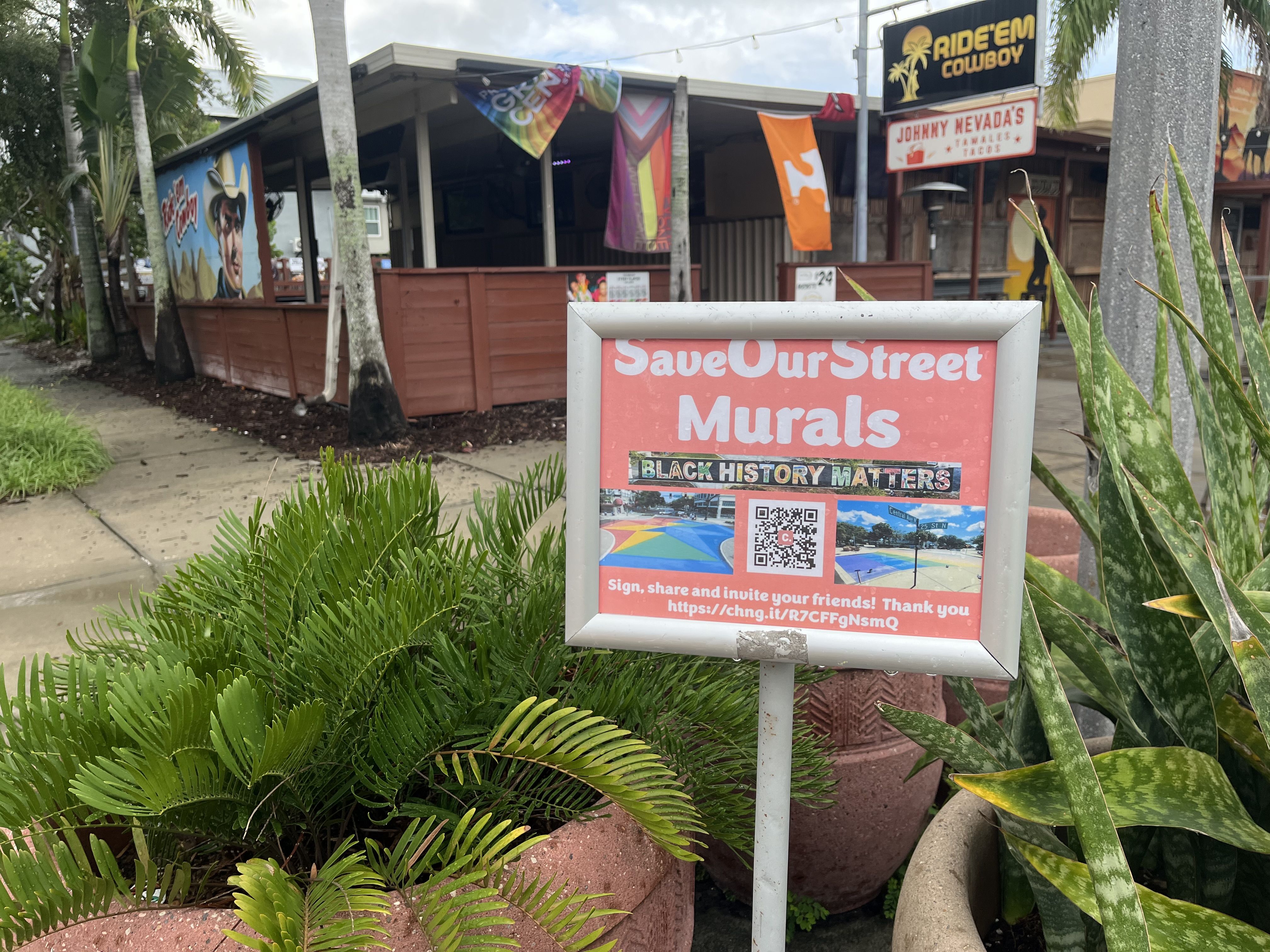 Outdoor sign advocating to save street murals, highlighting Black History Matters with images and a QR code, in front of a building with a cowboy mural and tropical plants.