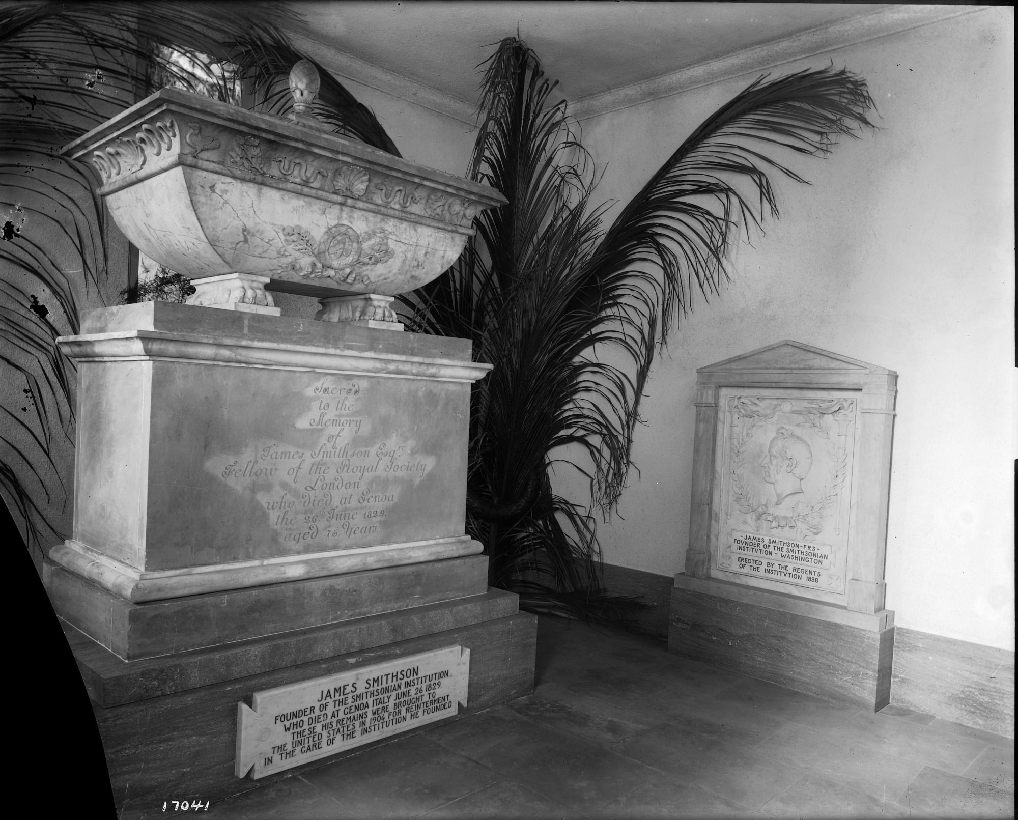 Black-and-white photo of a large marble urn on a stepped pedestal with carved reliefs, flanked by palm fronds; nearby plaques mention James Smithson, founder of the Smithsonian Institution.