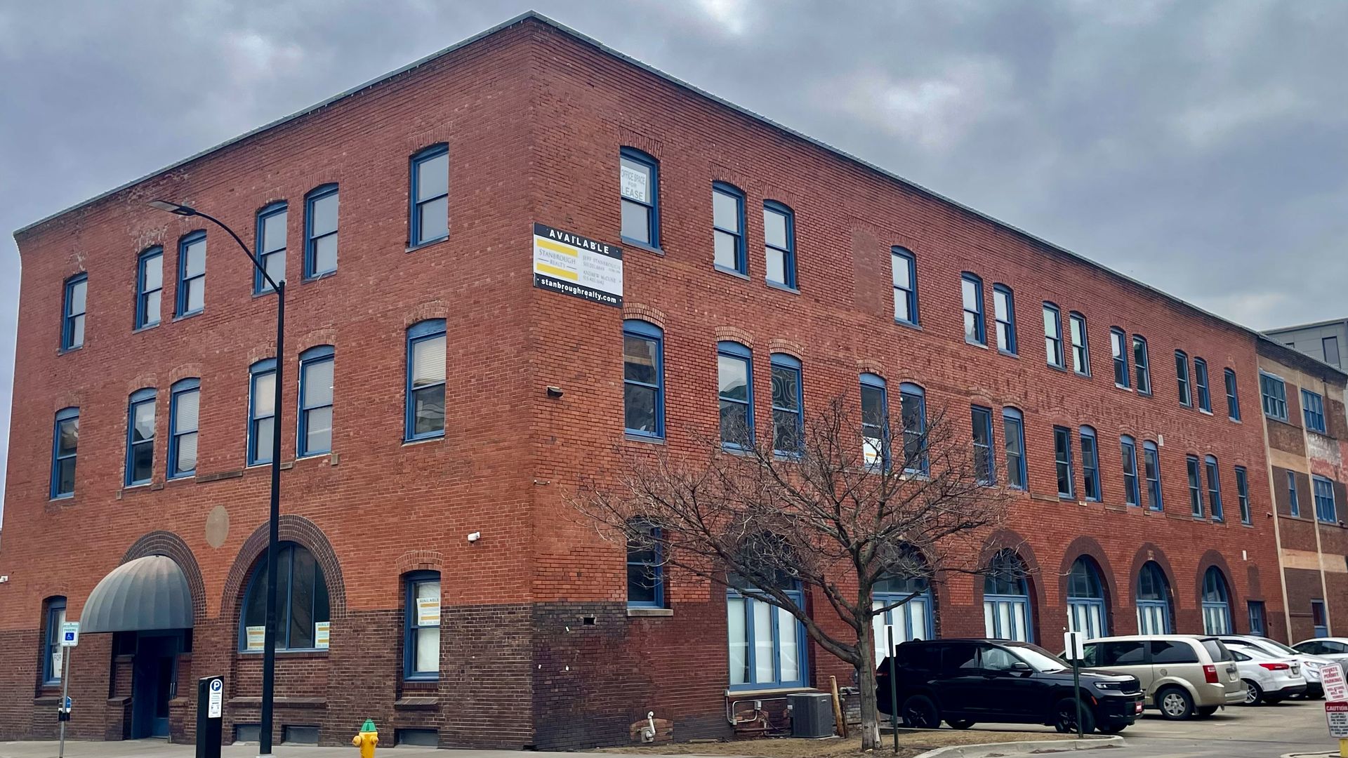 Three-story red brick building with blue-trimmed windows and arched entrances, a leafless tree in front, parked cars, and cloudy sky above.