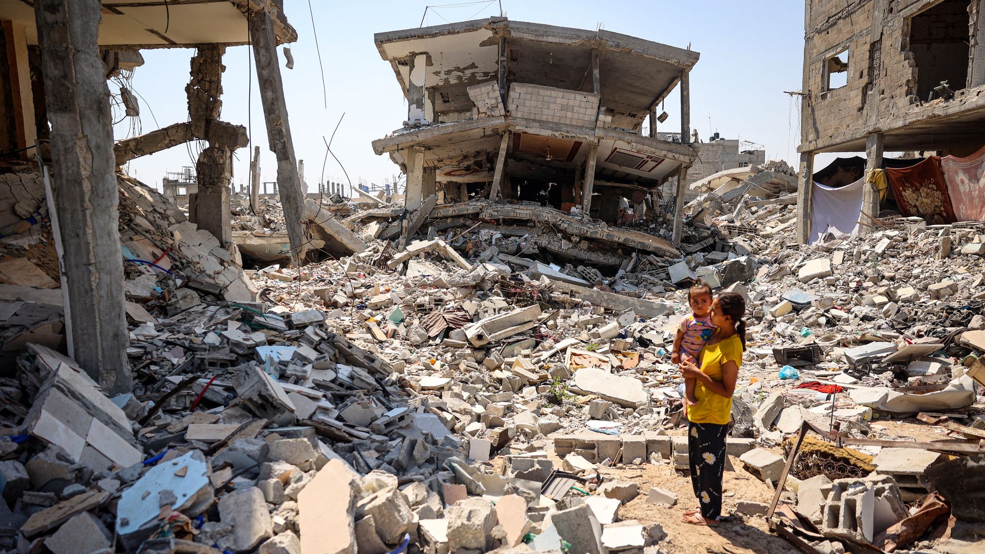 A woman stands holding a child surrounded by the rubble of buildings destroyed during Israeli bombardment in Khan Yunis on the southern Gaza Strip on June 23, 2024,