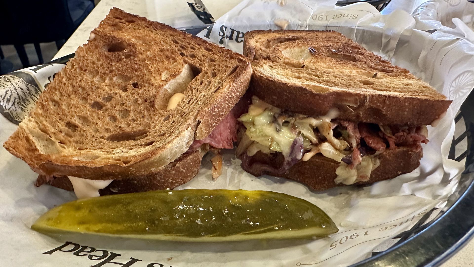 Two halves of a toasted sandwich with rye bread, melted cheese, and meat, served with a green pickle spear on a paper-lined metal tray.