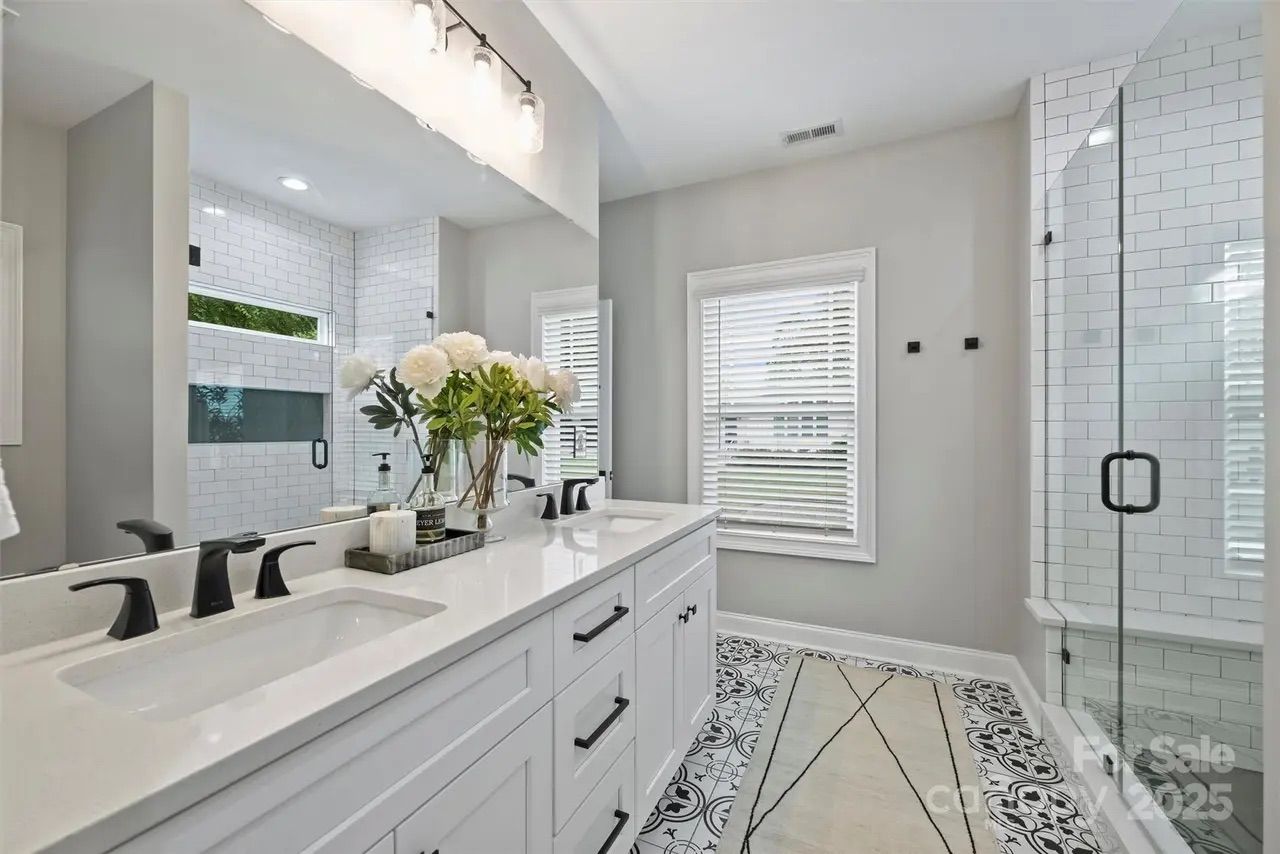 Bright bathroom with white double-sink vanity, black faucets, floral arrangement, patterned black and white floor tiles, walk-in glass shower, and window with white blinds.