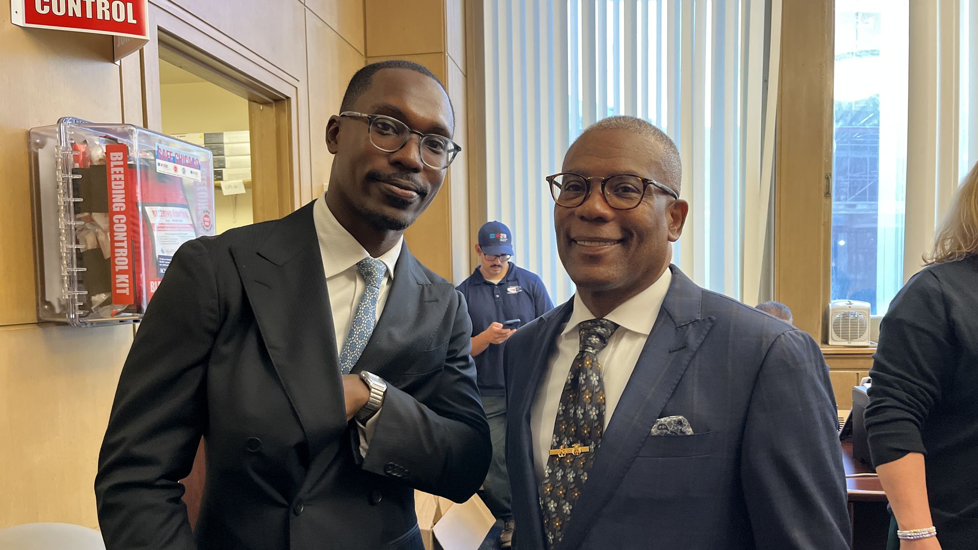 Two men in suits and glasses standing indoors near a bleeding control kit, one wearing a black suit and blue tie, the other in a navy suit with a floral tie and pocket square.